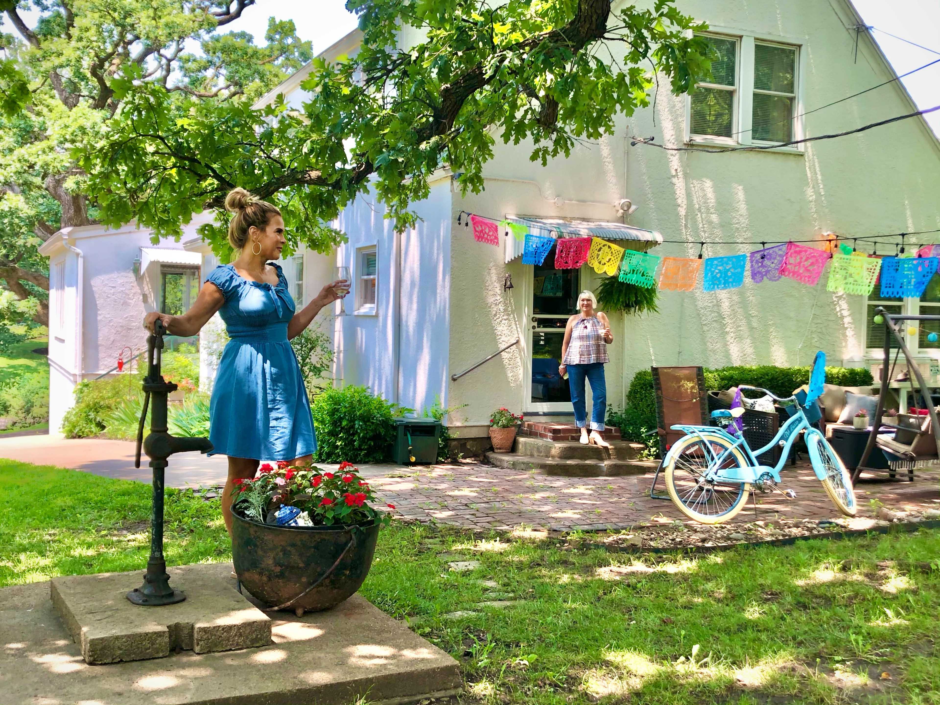 A woman in a blue dress stands by a garden planter while another woman holds a drink on a porch, with colorful decorations hanging in the background near a house.