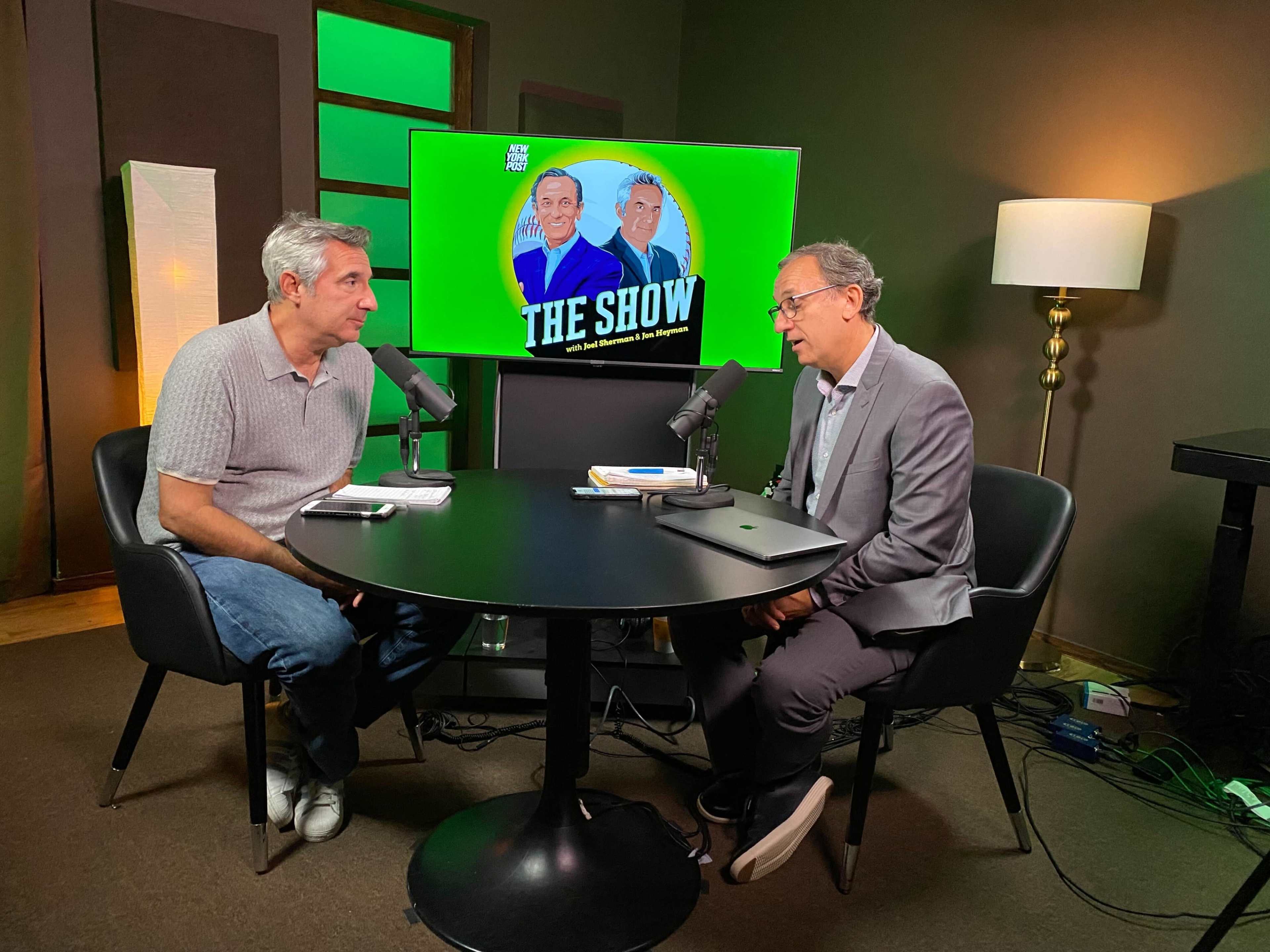 Two men are sitting at a round table with microphones in front of them, discussing in a studio setting with a green backdrop displaying "THE SHOW."