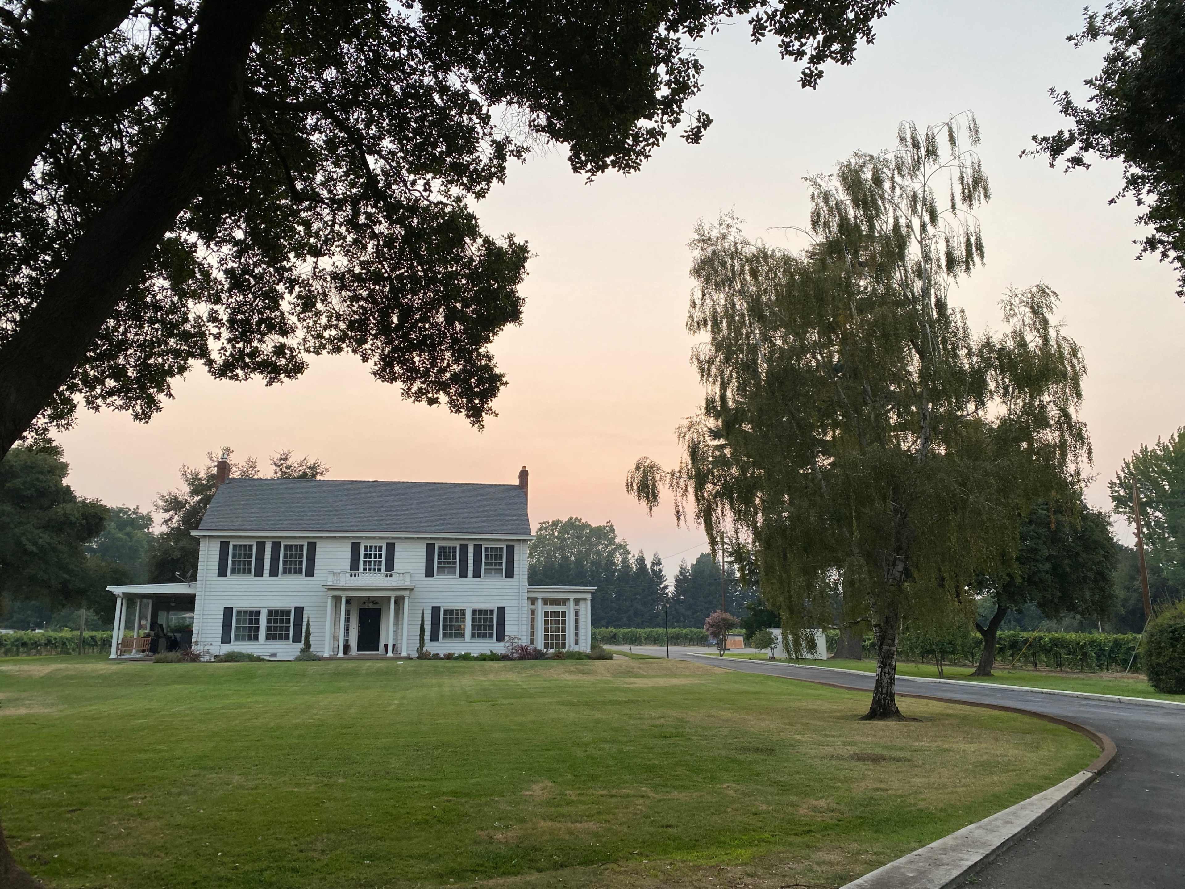 A large white house with a gray roof is set on a grassy area, surrounded by trees and near a winding road.
