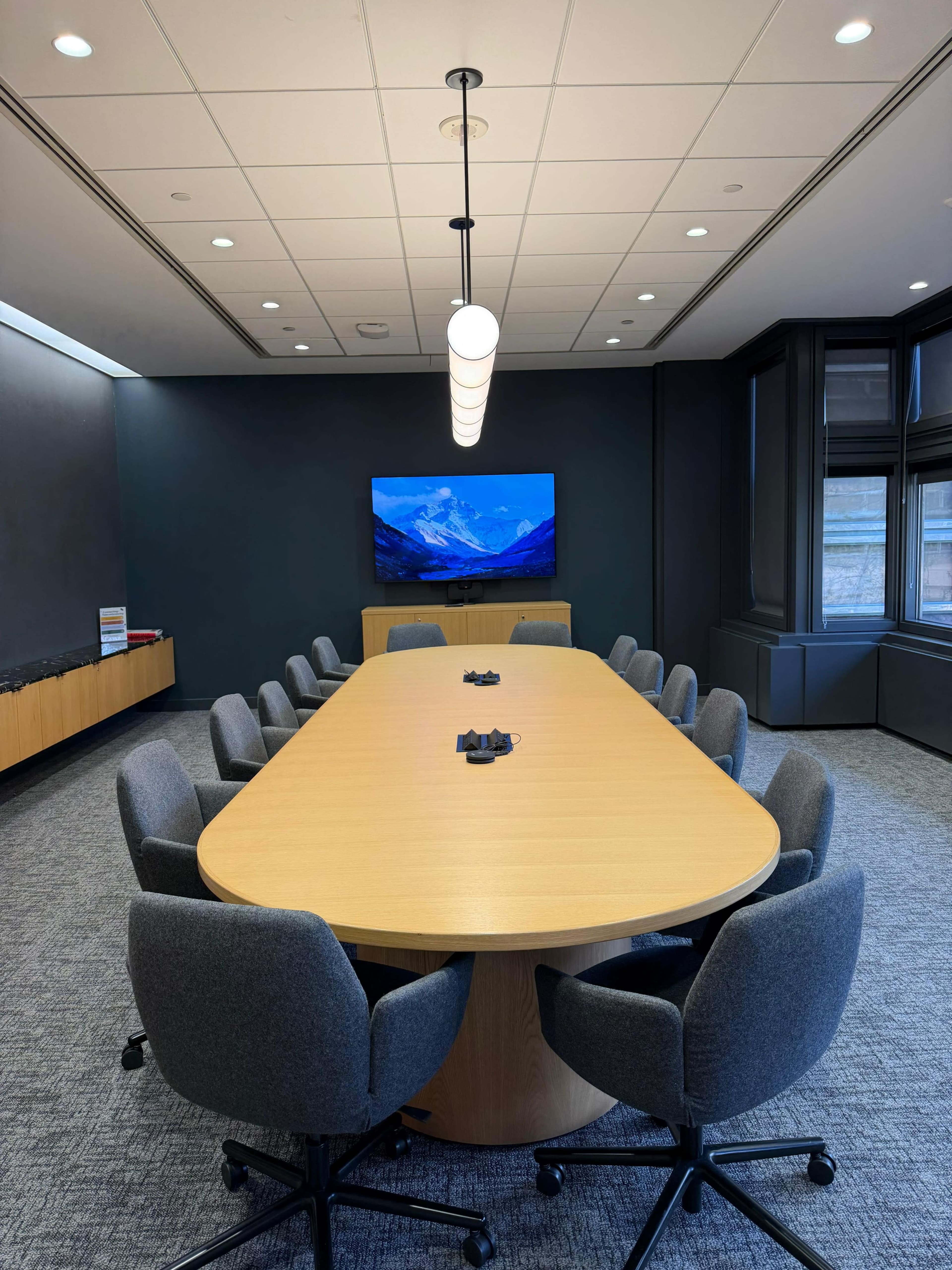 The image shows a modern conference room featuring a long oval table surrounded by ergonomic chairs, with a large screen displaying a mountain landscape on the wall.