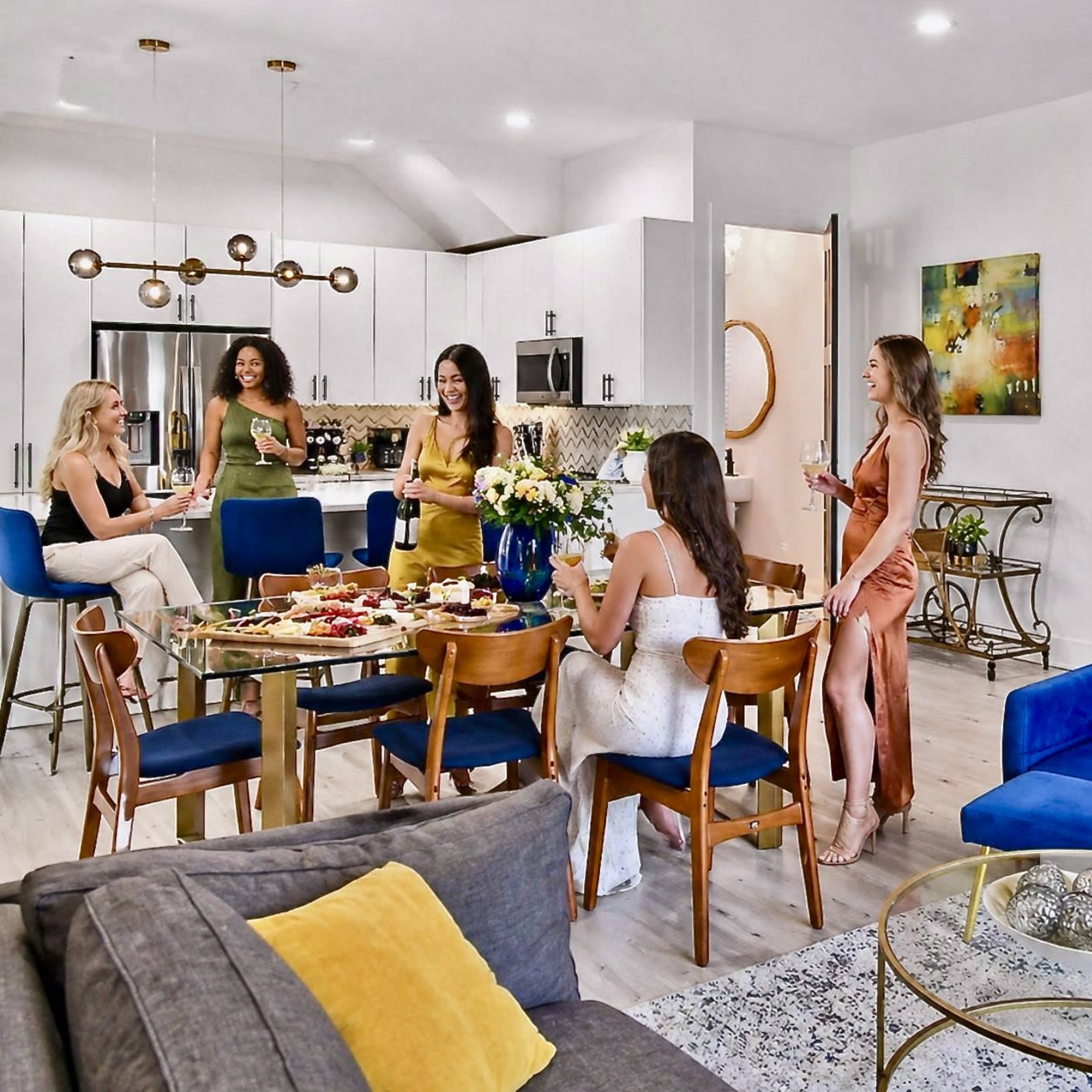 A group of five women is gathered around a dining table in a modern kitchen, sharing food and drinks.