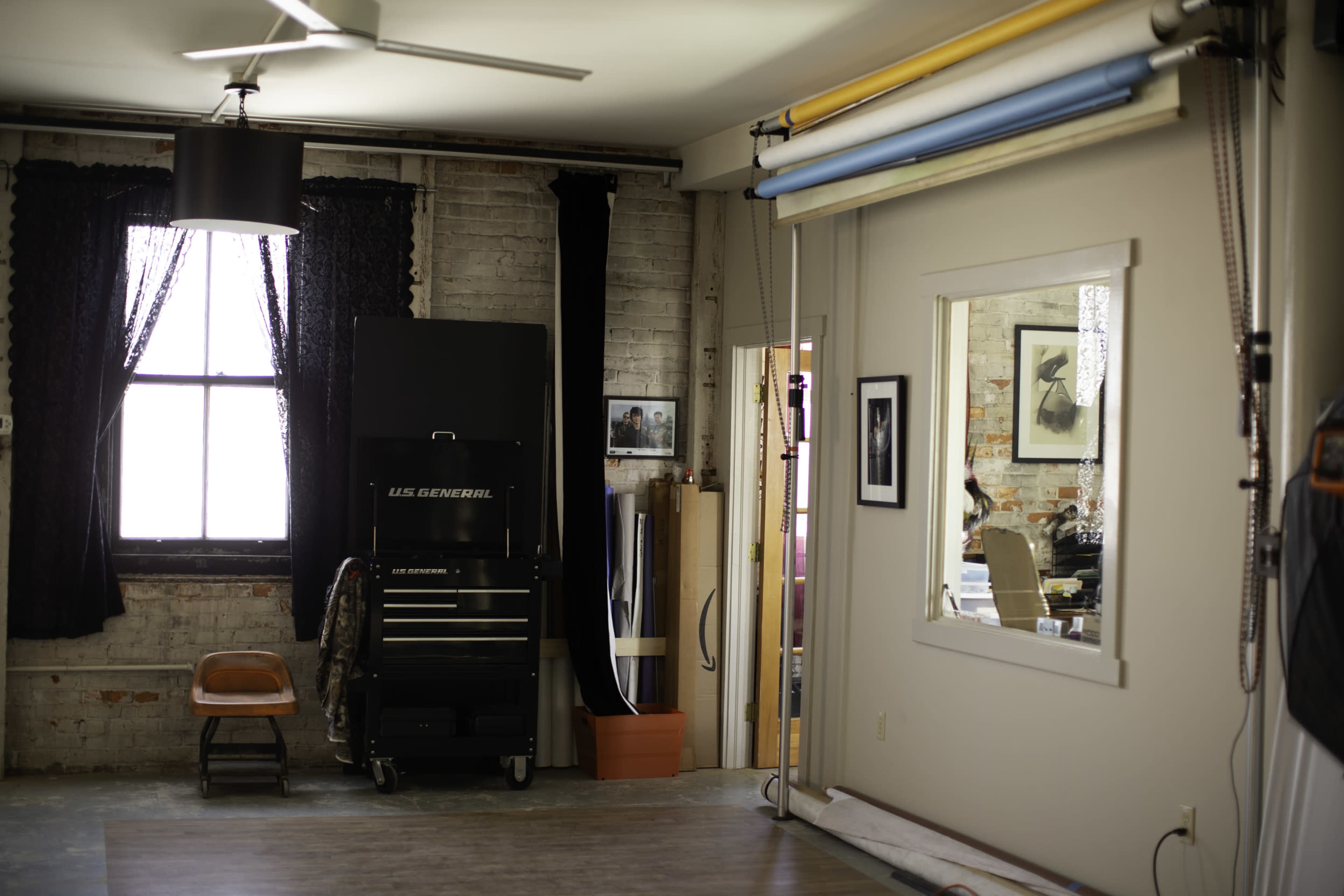 A well-lit studio space with a large window, a tool cabinet, a mirror, and different colored backdrop rolls hanging on the wall.