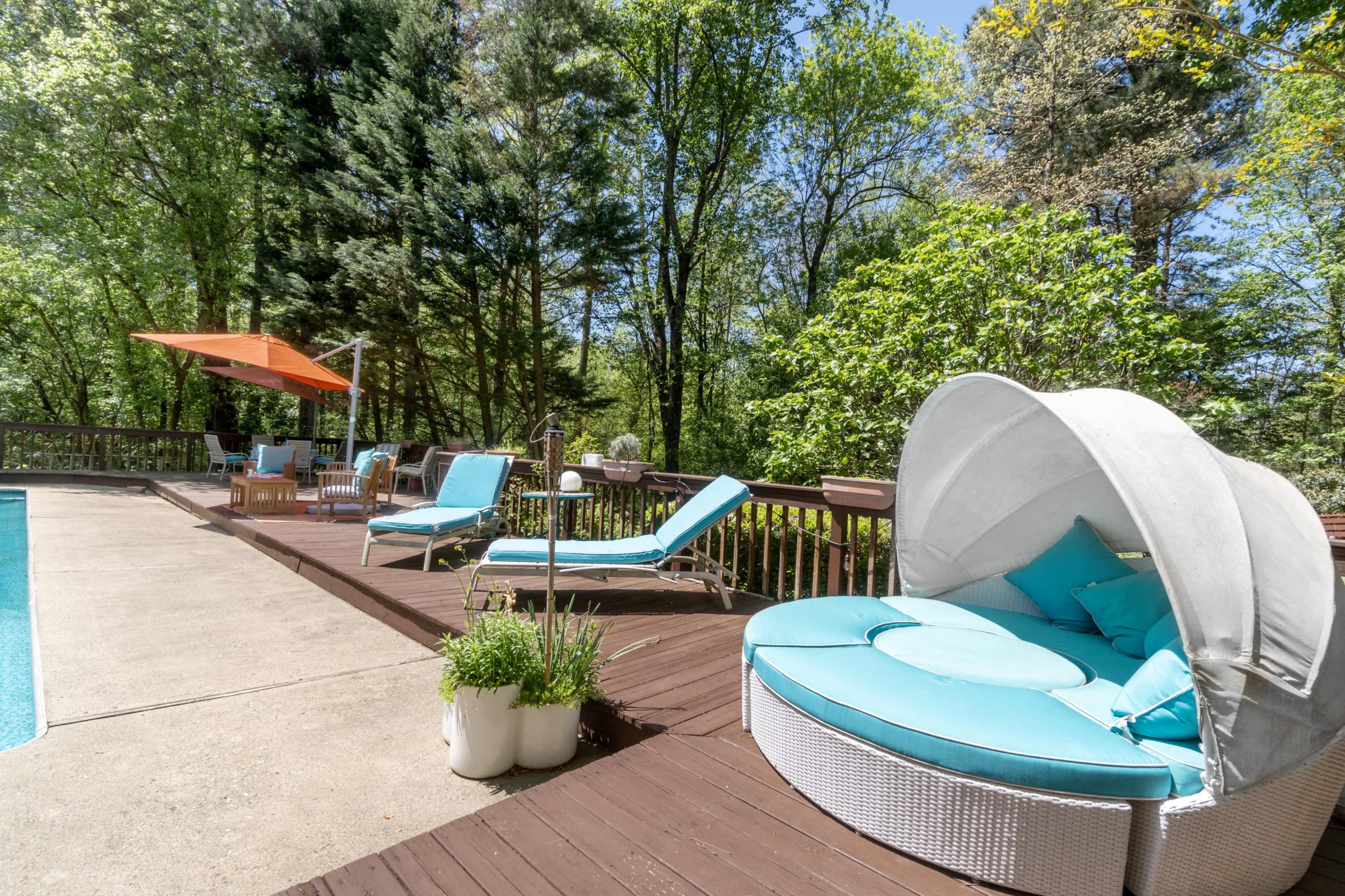 A poolside area featuring a round, cushioned lounge chair, two chaise lounges, and an orange umbrella, surrounded by lush greenery.