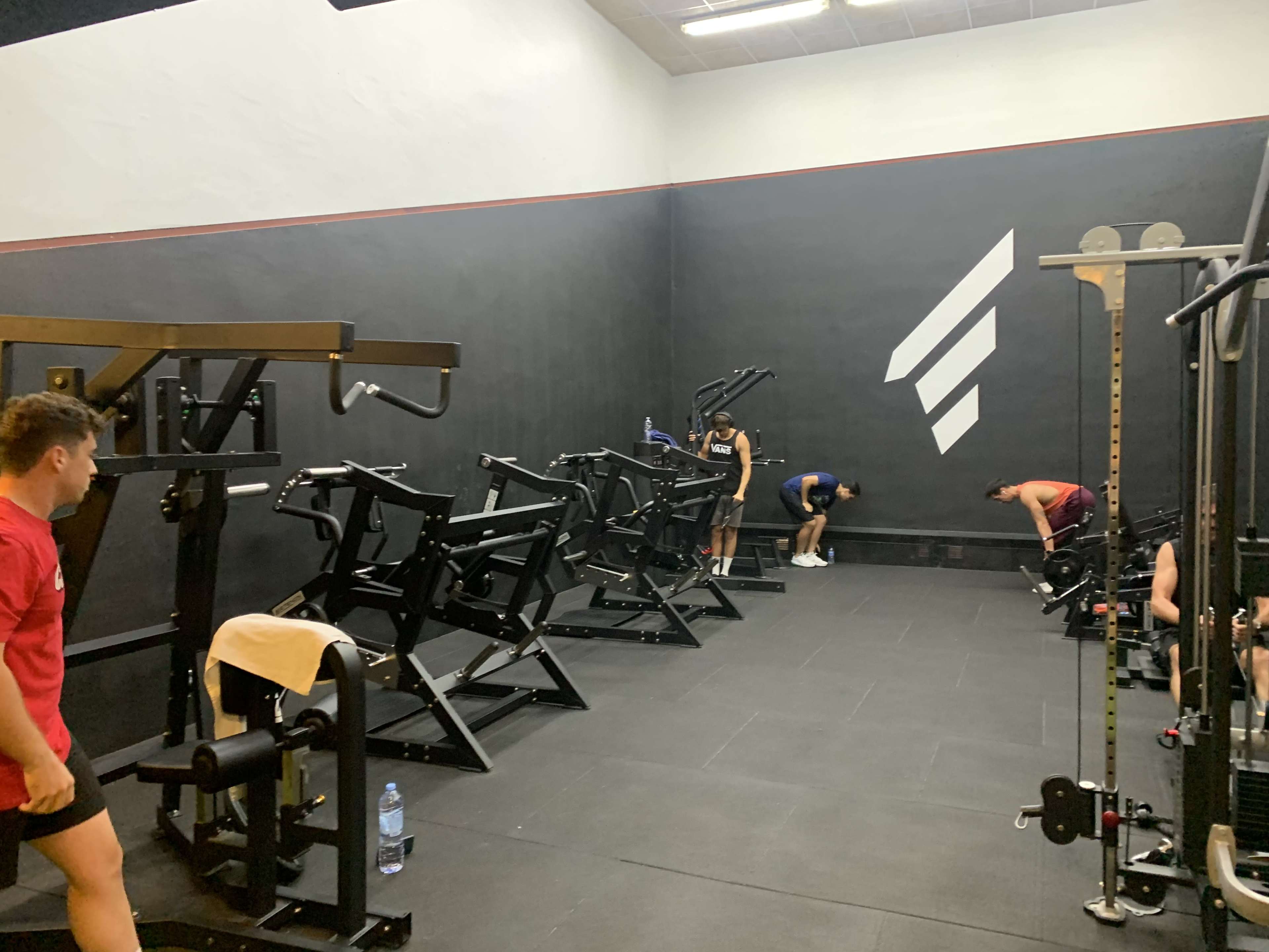 A dimly lit gym with workout equipment on one side and three individuals engaged in exercises.