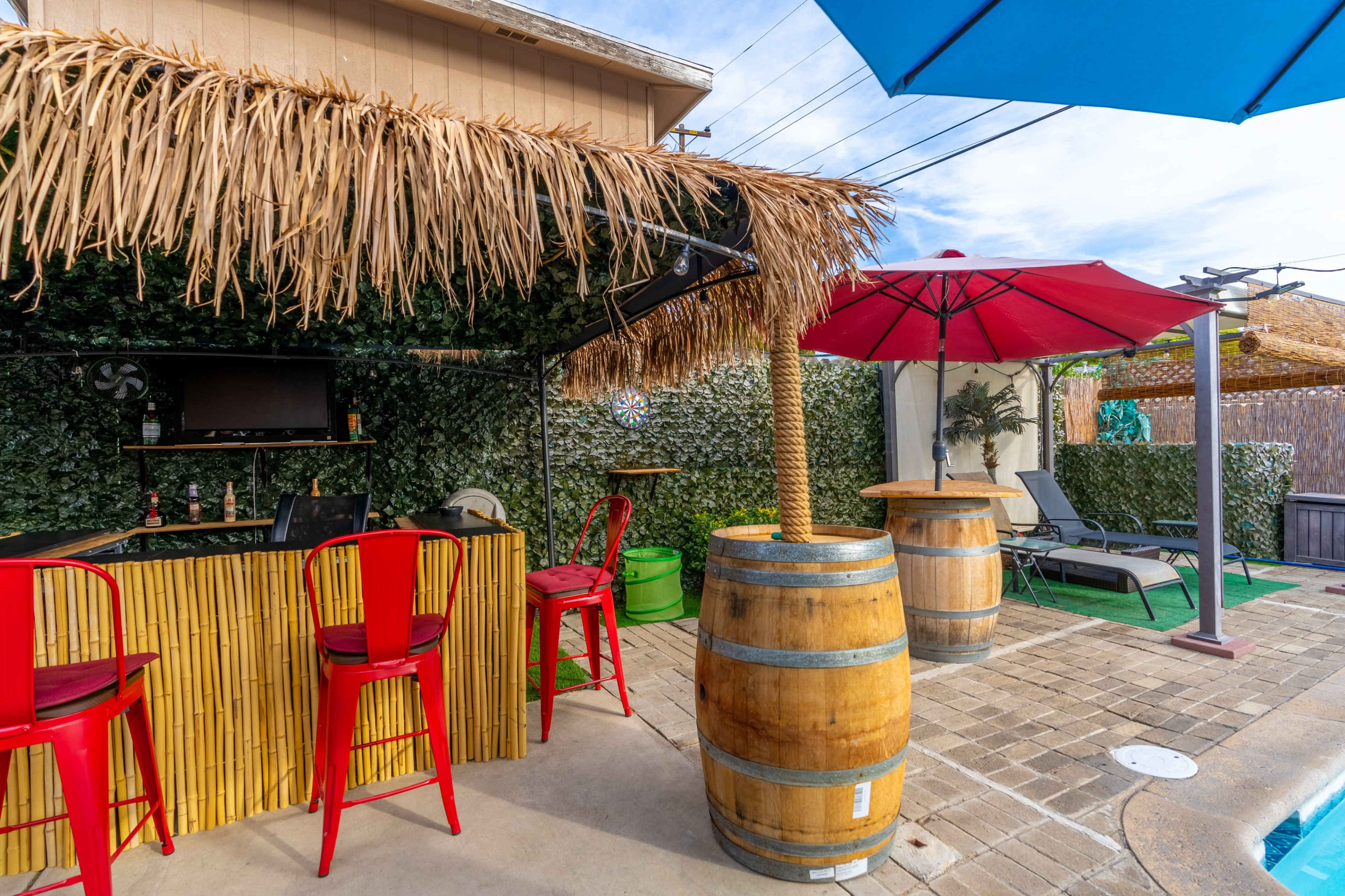 A tiki-style outdoor bar with bamboo accents, red stools, and a blue and red umbrella, surrounded by greenery and a pool area.