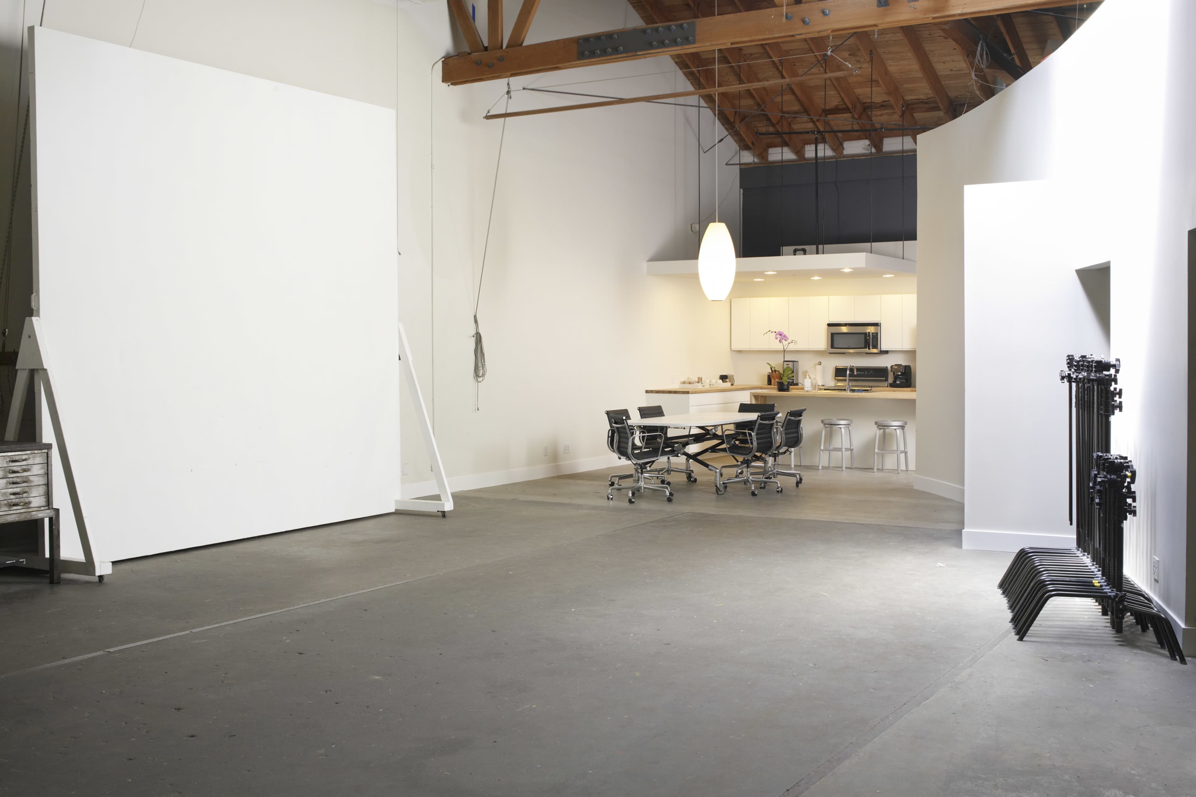 The image shows a spacious studio with a large white wall, a kitchen area in the background, and a meeting table surrounded by chairs.