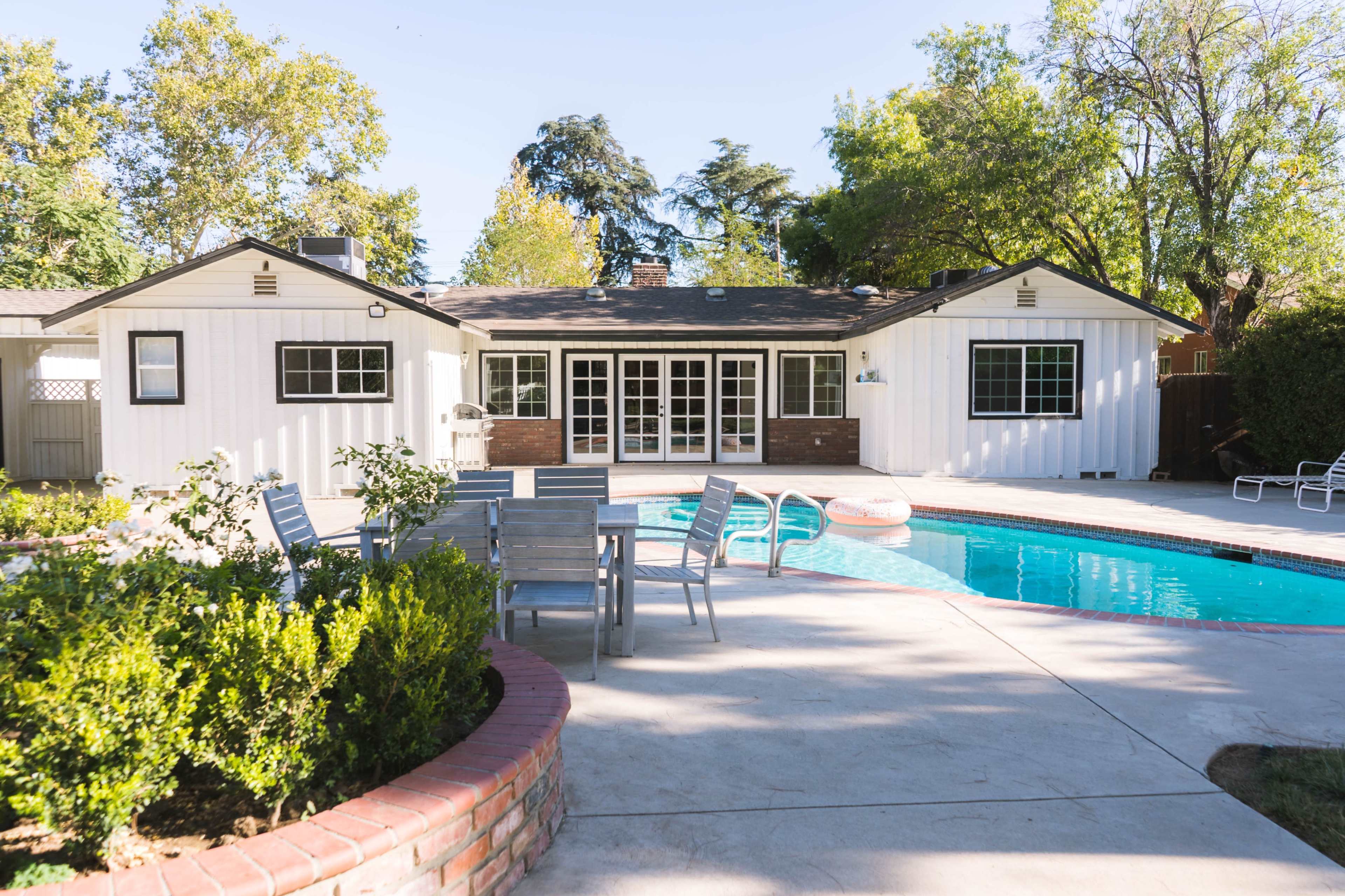 A backyard with a swimming pool surrounded by patio furniture and a single-story house in the background.