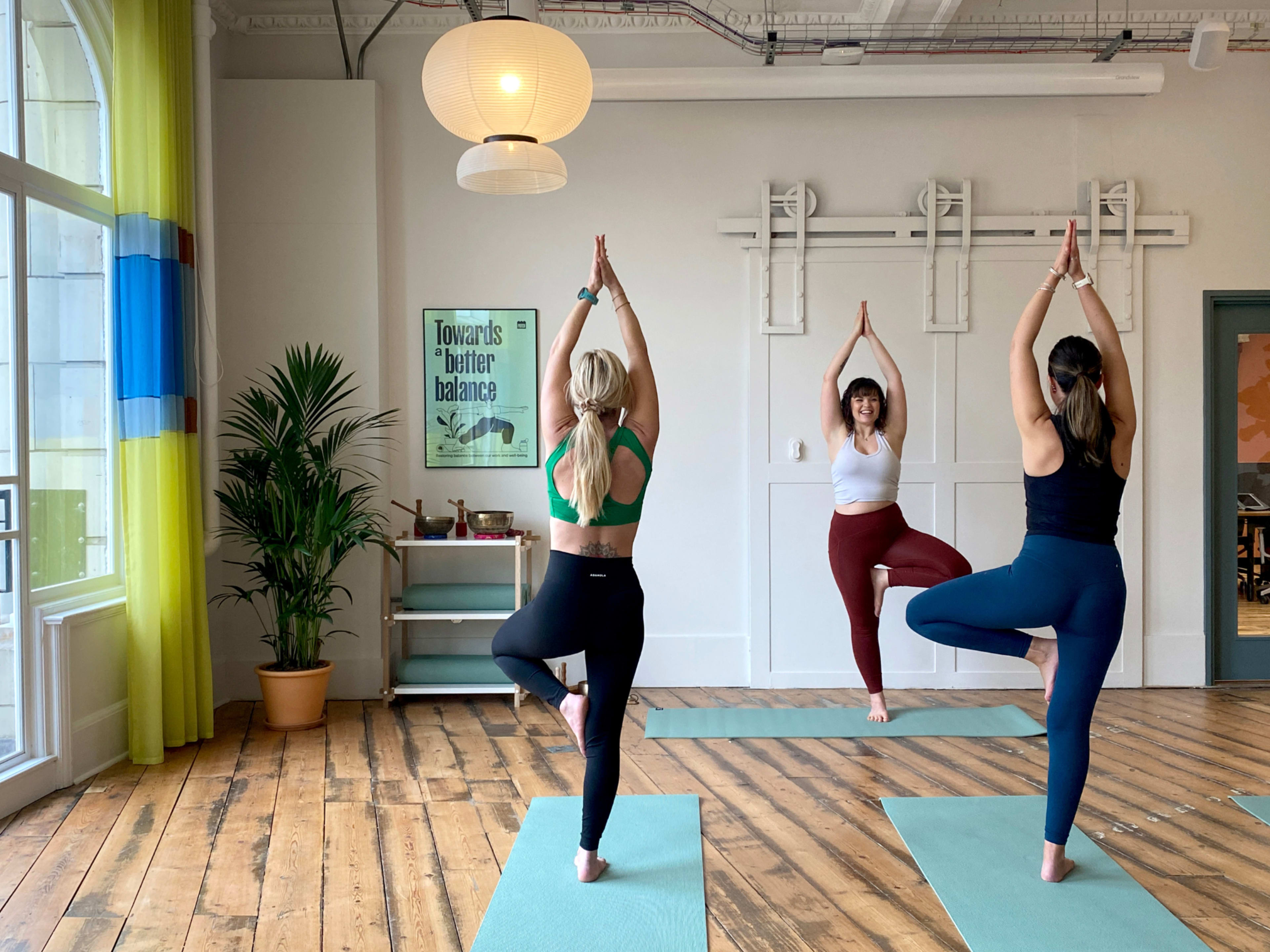 Three individuals are practicing yoga in a studio, standing on yoga mats in a tree pose with a plant and a light fixture visible in the background.