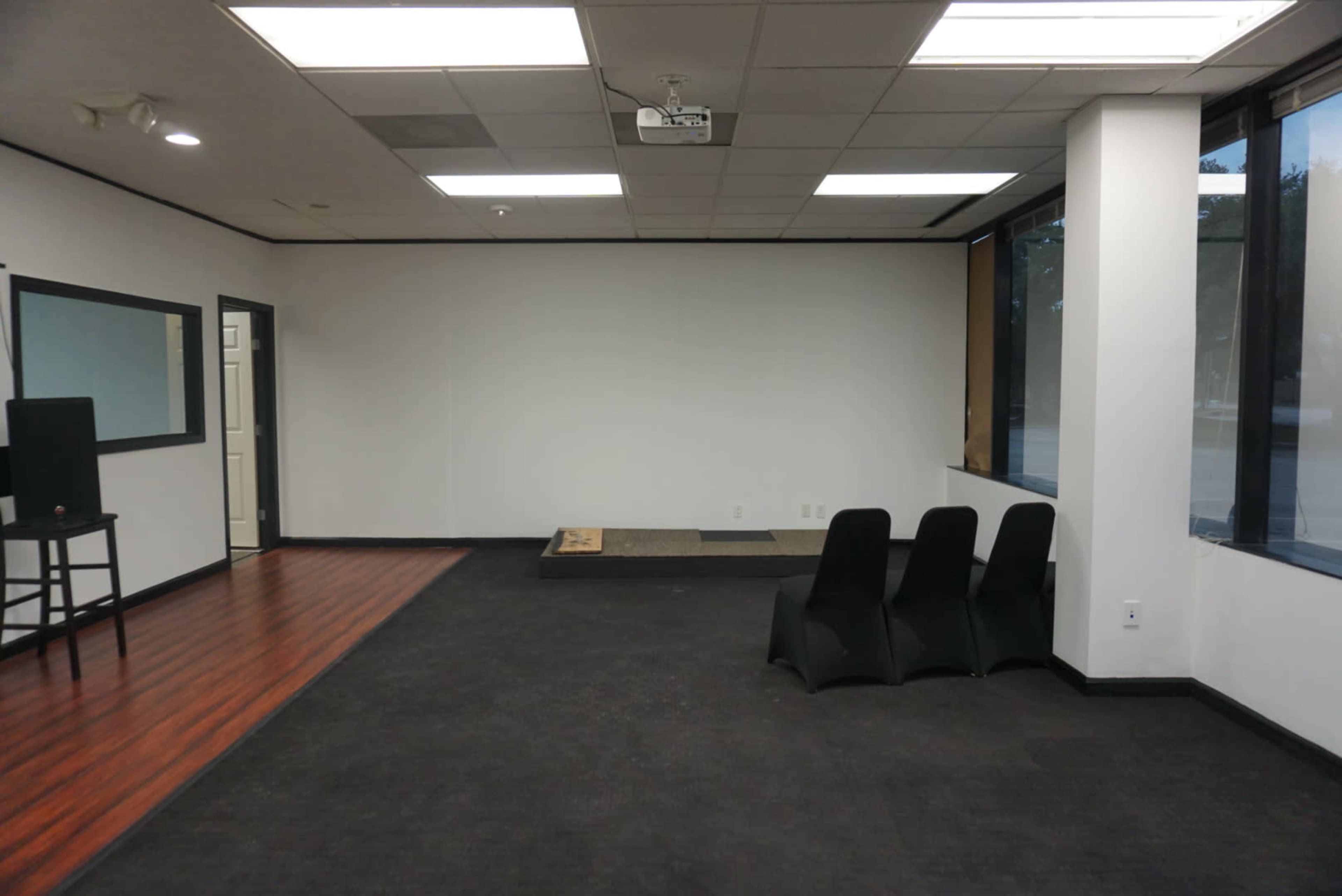 A sparsely furnished room with a dark floor, a small stage area, a projector mounted on the ceiling, and a few black chairs aligned against one wall.