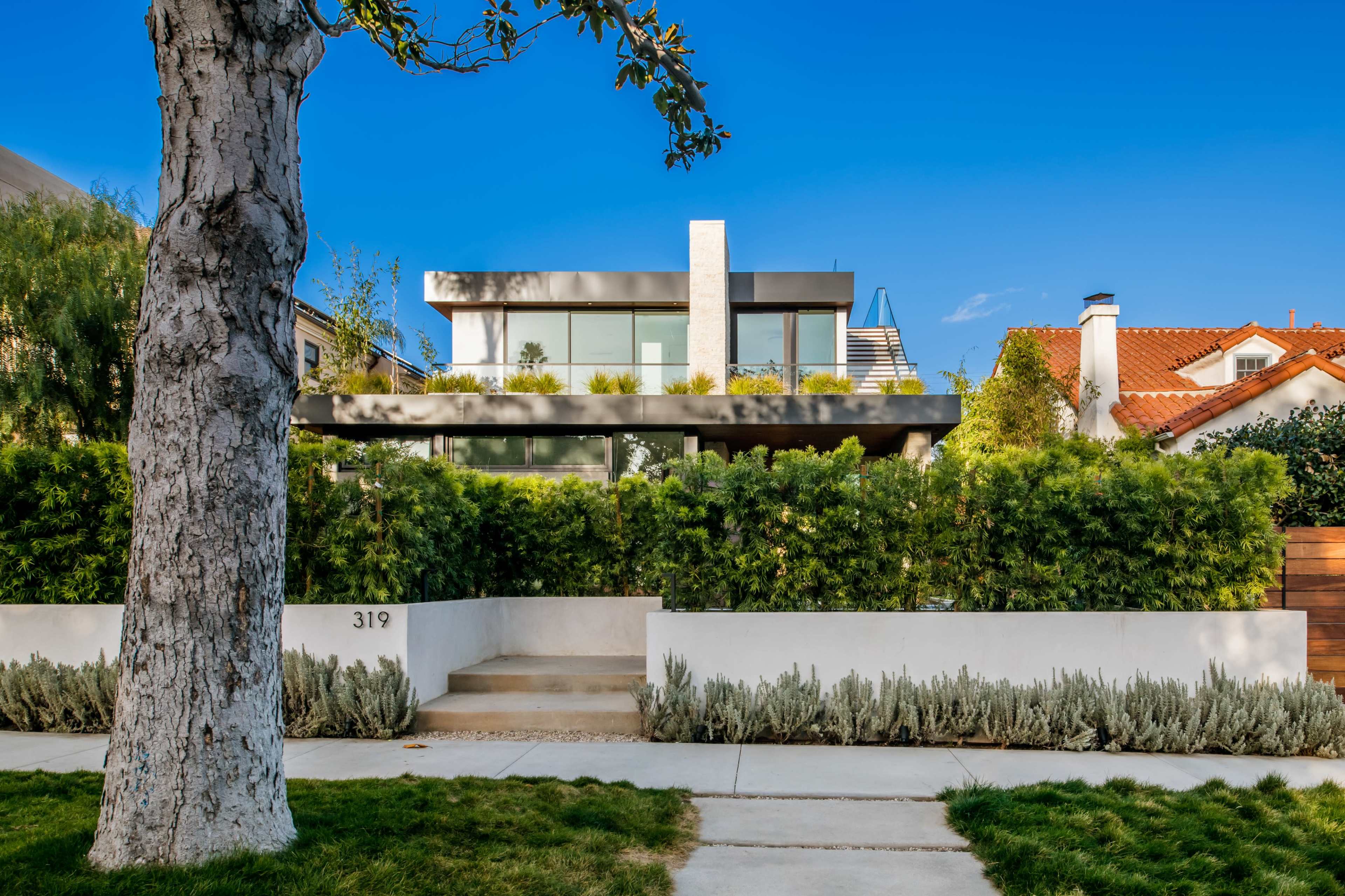 A modern two-story house with large windows is surrounded by a low hedge and a landscaped front yard.