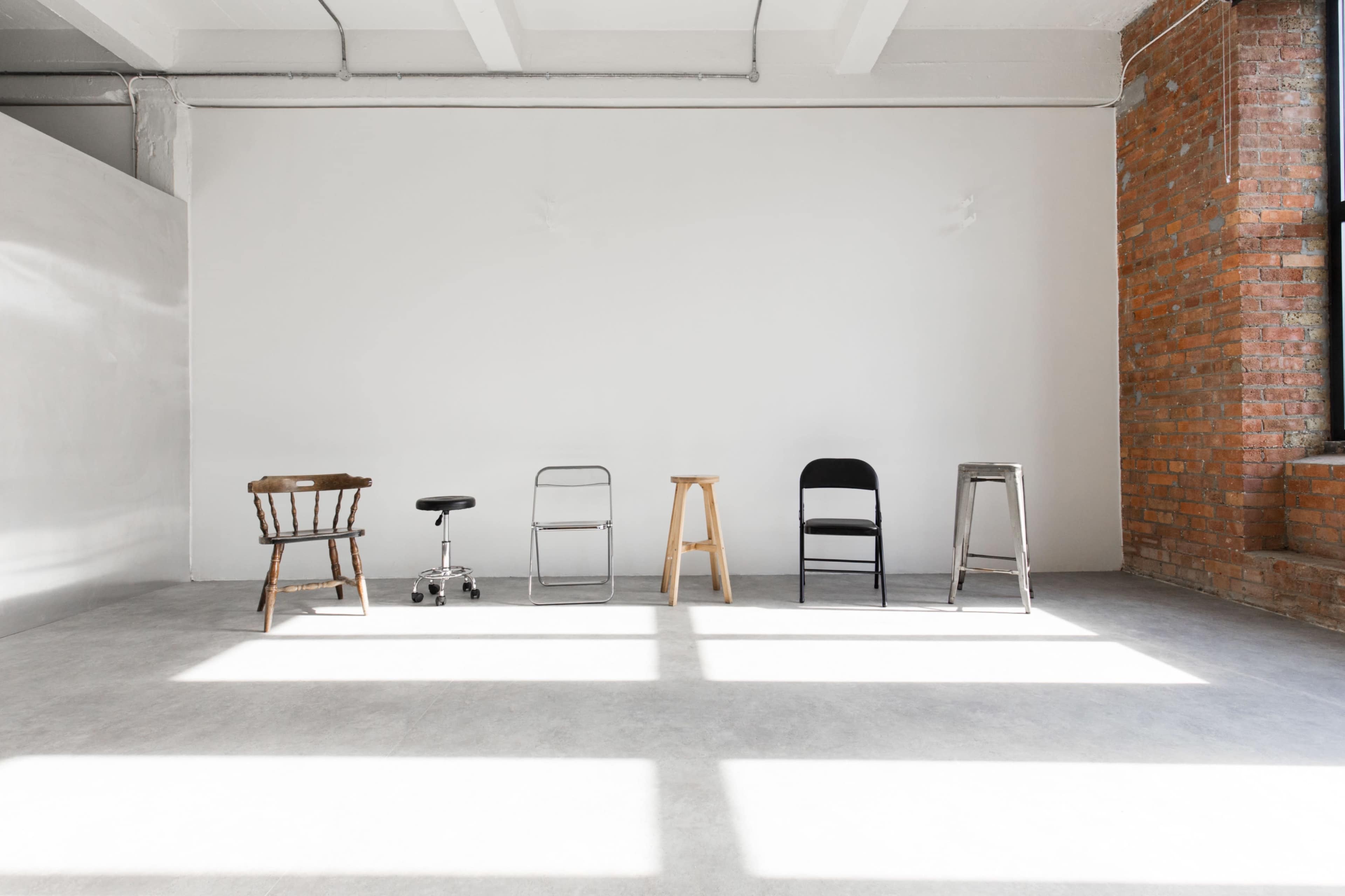 The image shows a row of five different types of stools arranged on a concrete floor in a brightly lit room with a plain white wall and exposed brick.