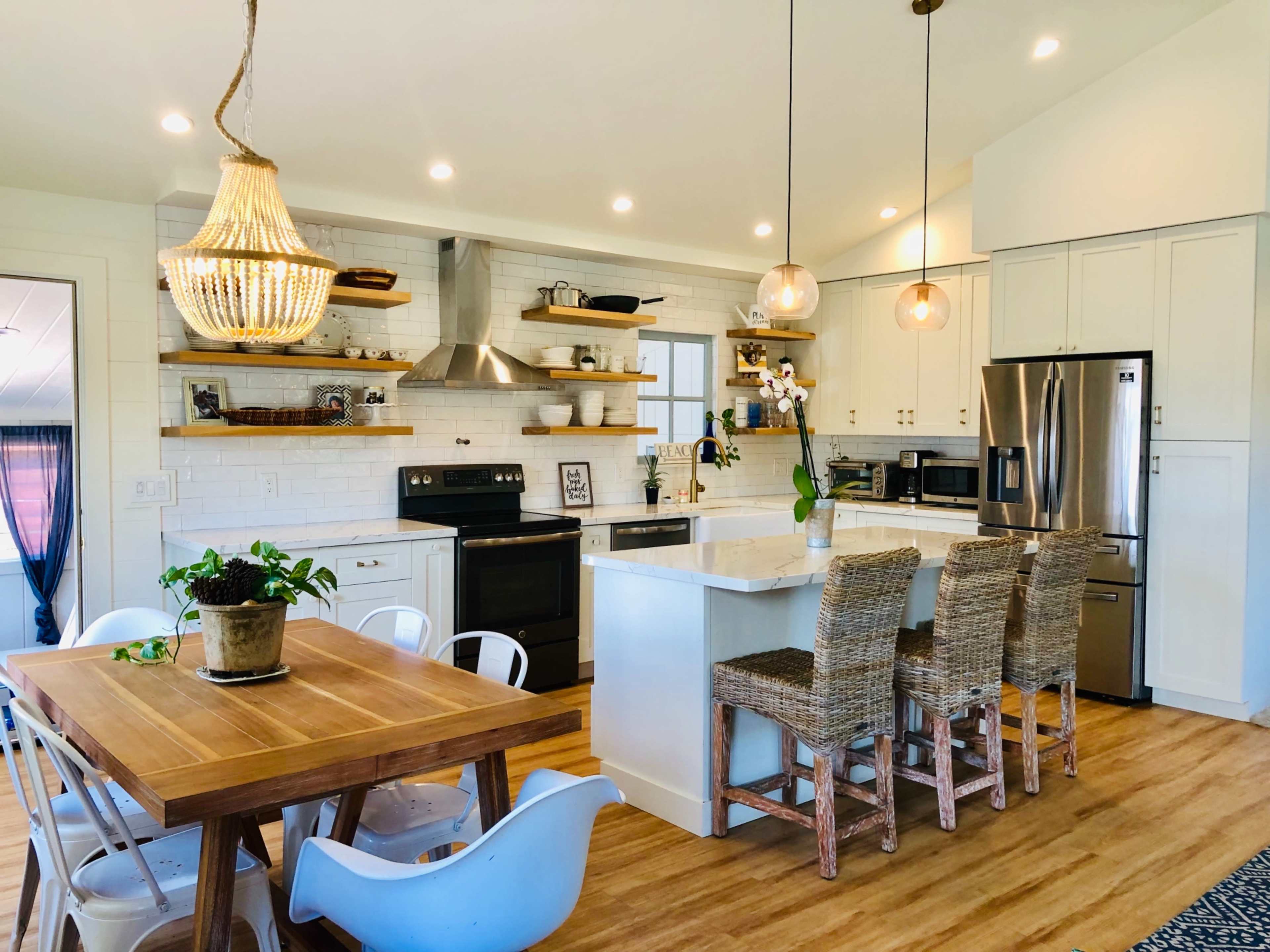 A modern kitchen features white cabinetry, open shelving with decorative items, a large island with seating, and a wooden dining table.