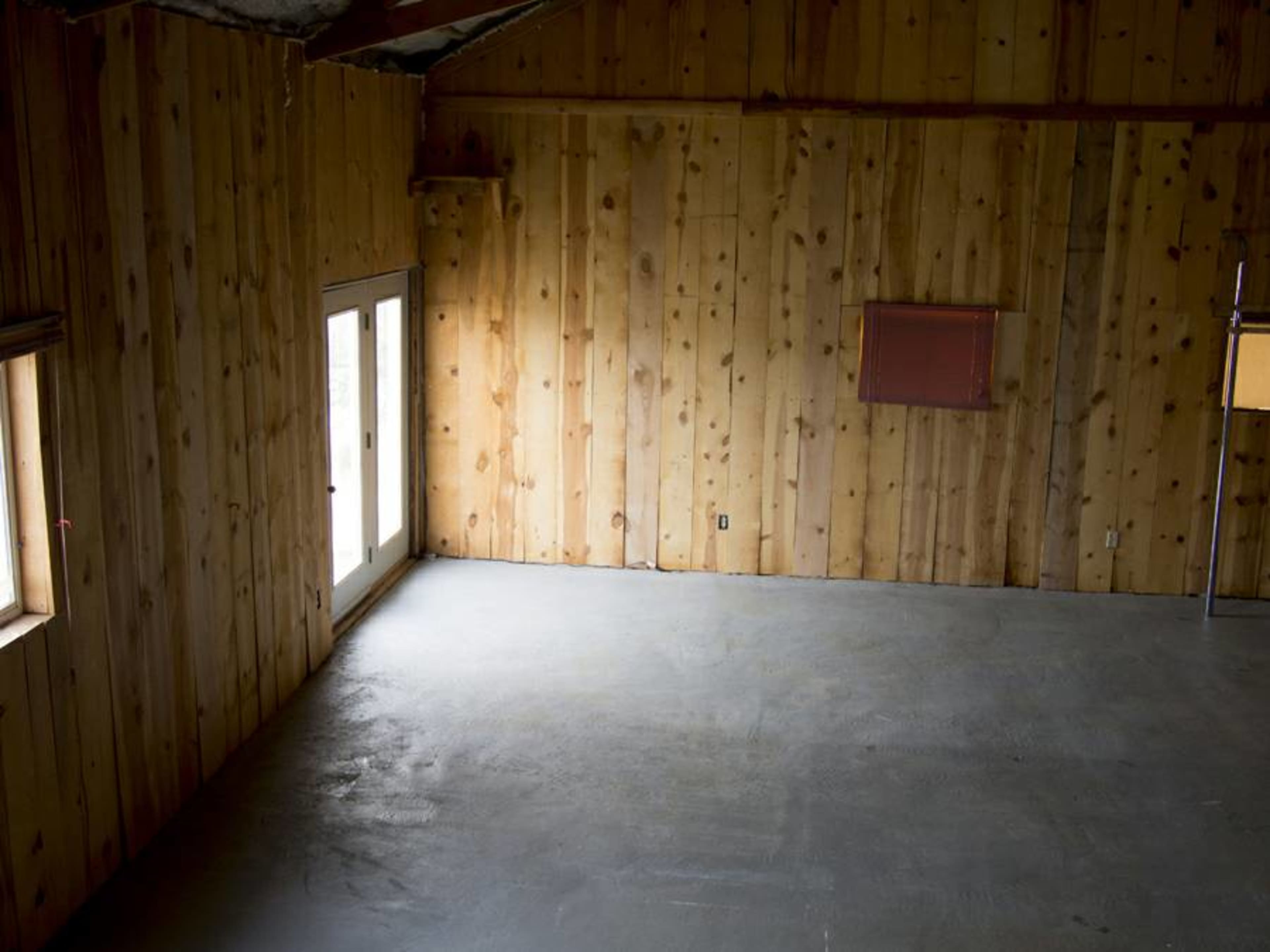 The image shows an empty room with wooden walls, a concrete floor, and a window on one side.