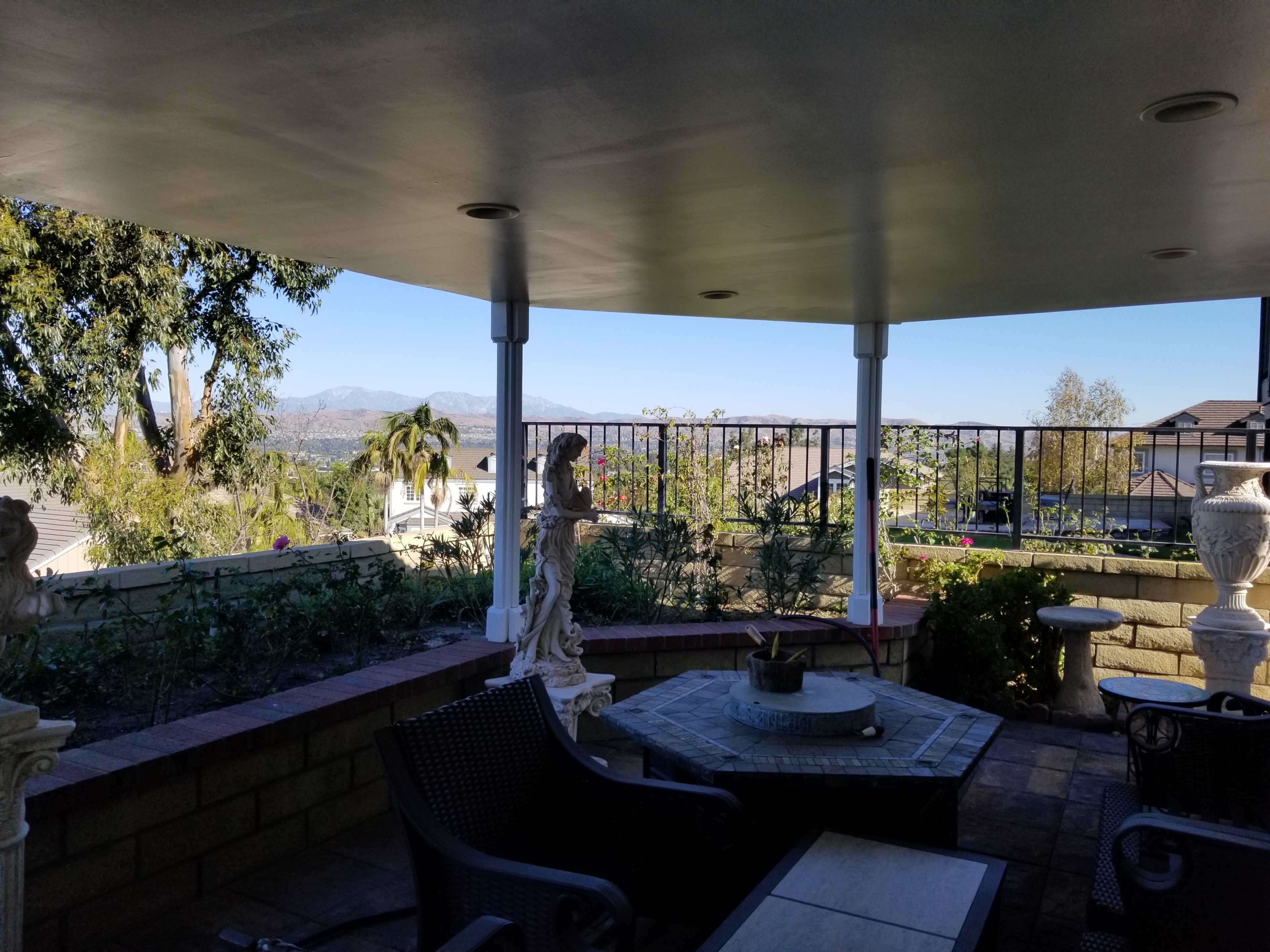 The image shows a covered patio area with outdoor furniture, overlooking a landscaped yard and distant mountains.