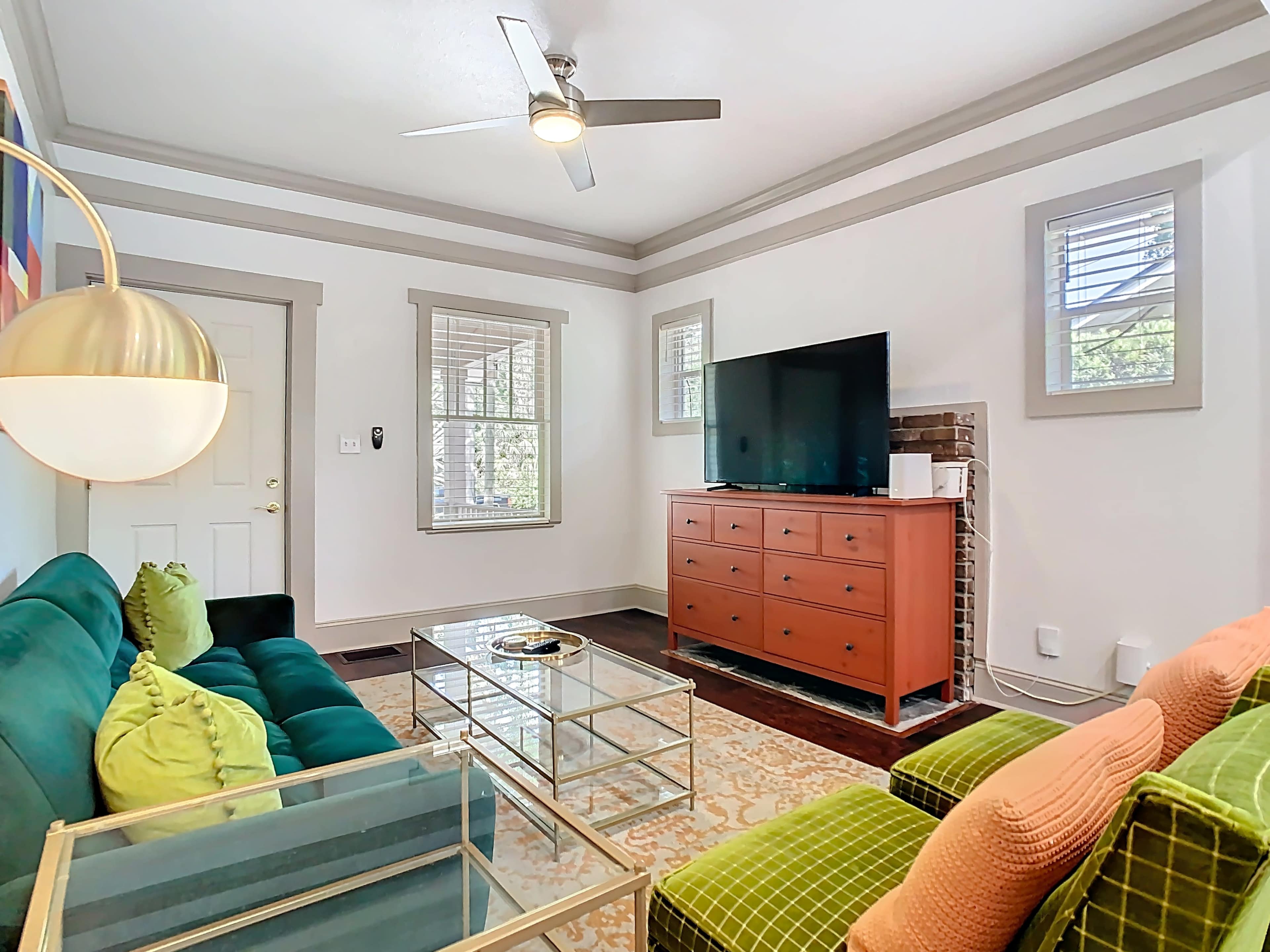 The living room features a green couch and a tan loveseat, a glass coffee table, a wooden television cabinet, and large windows allowing natural light to enter.
