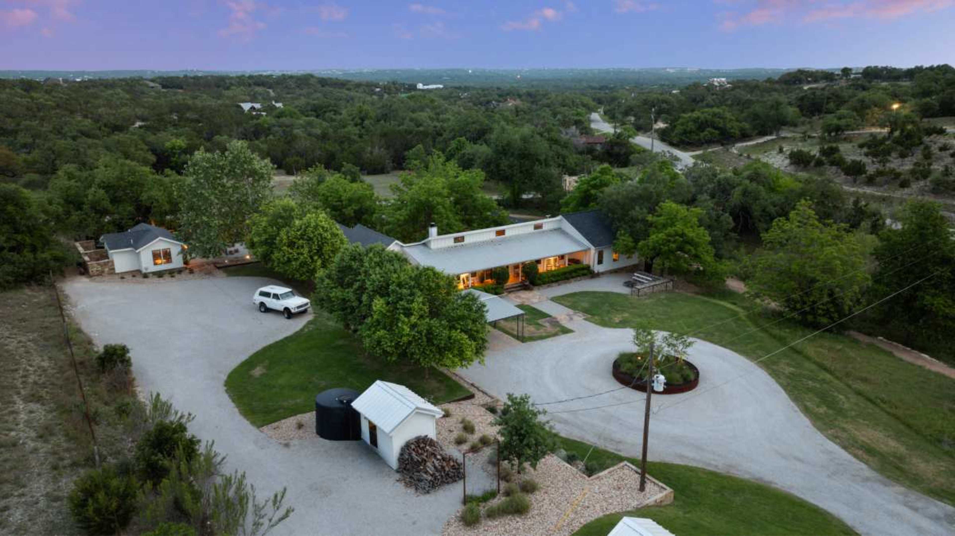The image shows a spacious, rural property with a large house surrounded by greenery, a winding driveway, and various outbuildings.