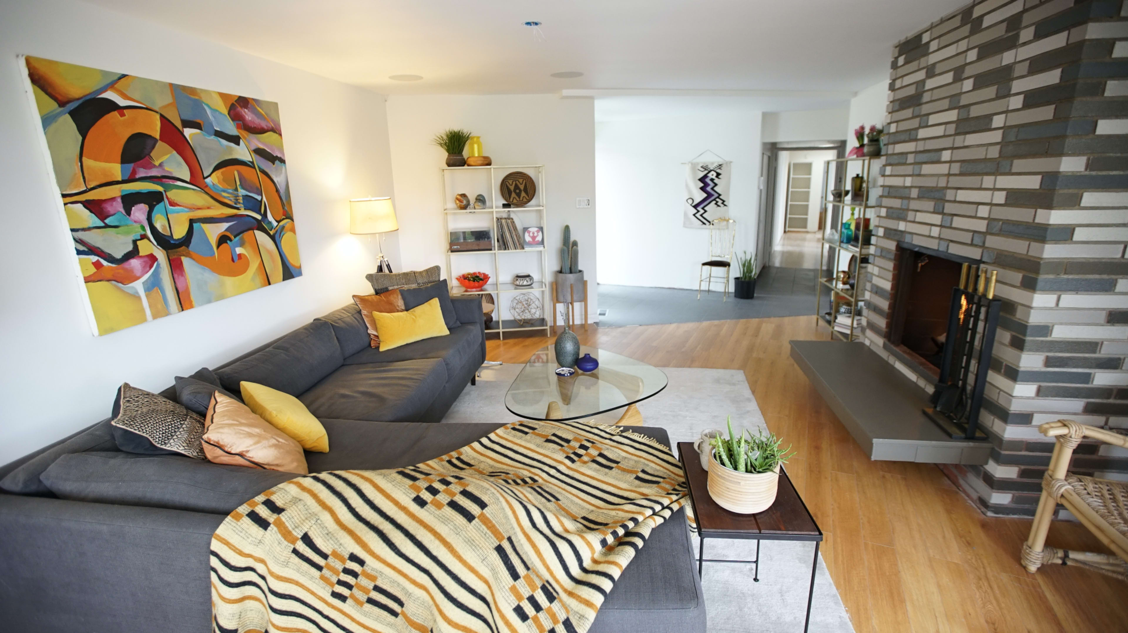 A modern living room features a gray sectional sofa with colorful cushions, a glass coffee table, and a textured wall adorned with an abstract painting and a stone fireplace.
