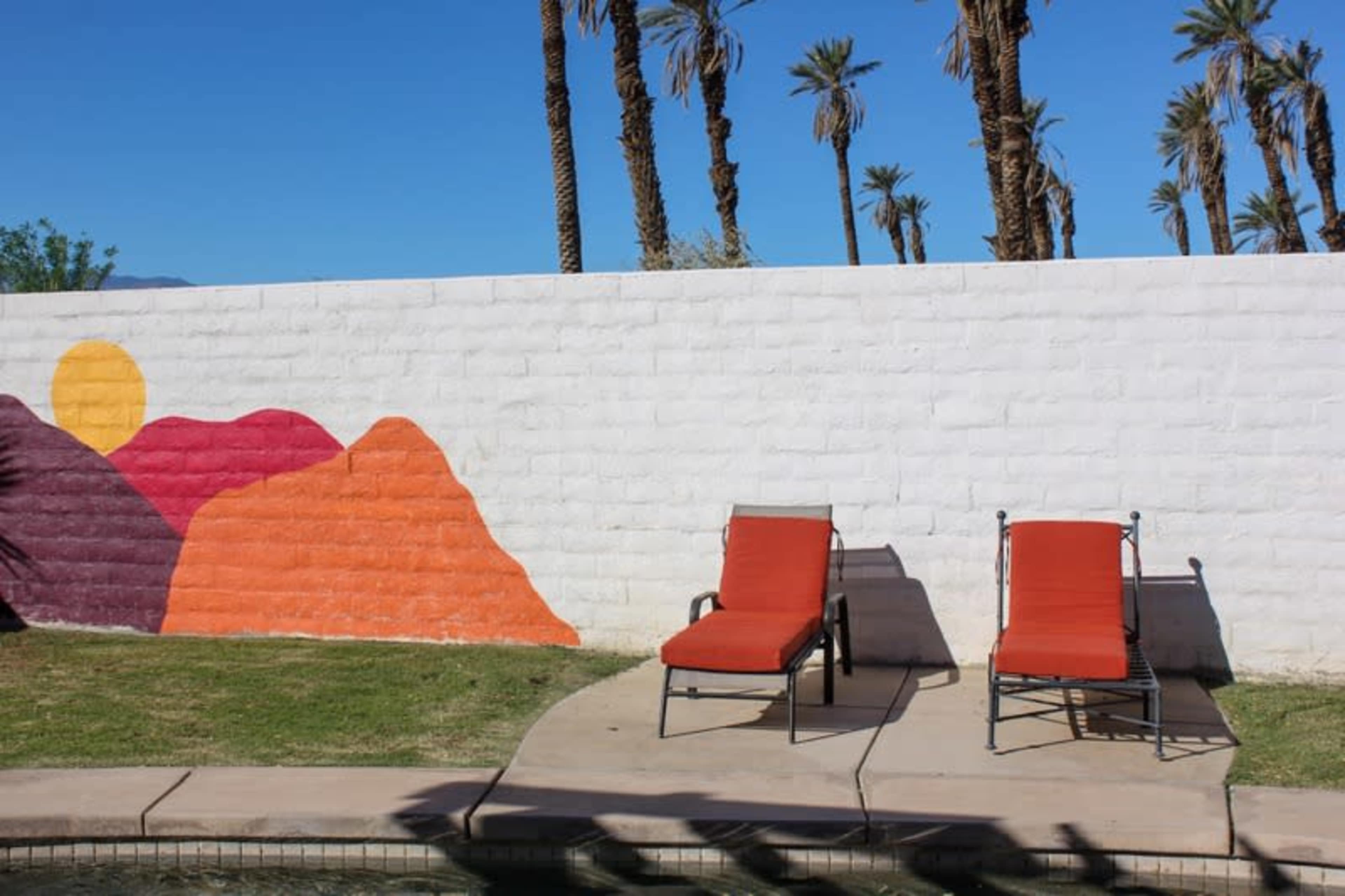 Two red lounge chairs sit on a concrete patio beside a swimming pool, against a white wall decorated with a colorful mountain mural and framed by tall palm trees.