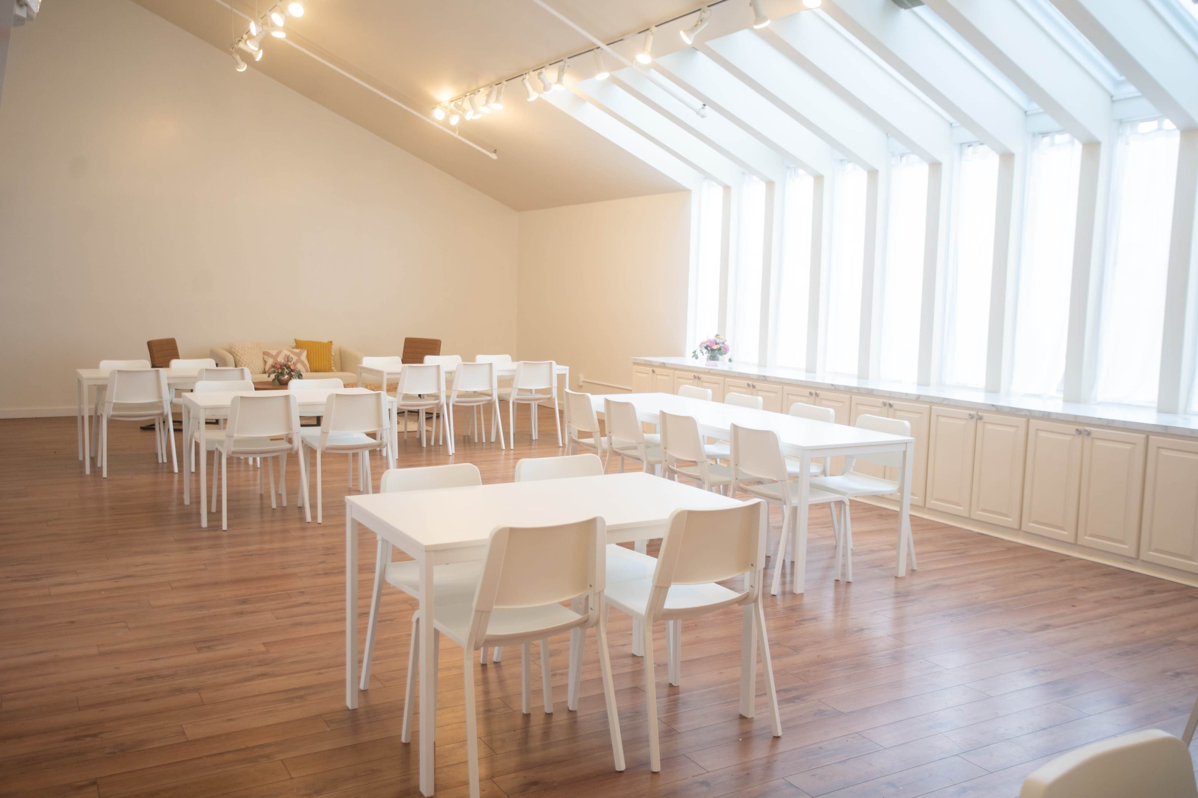 The image shows a bright, spacious room with several white tables and chairs arranged throughout, large windows along one wall, and polished wooden flooring.