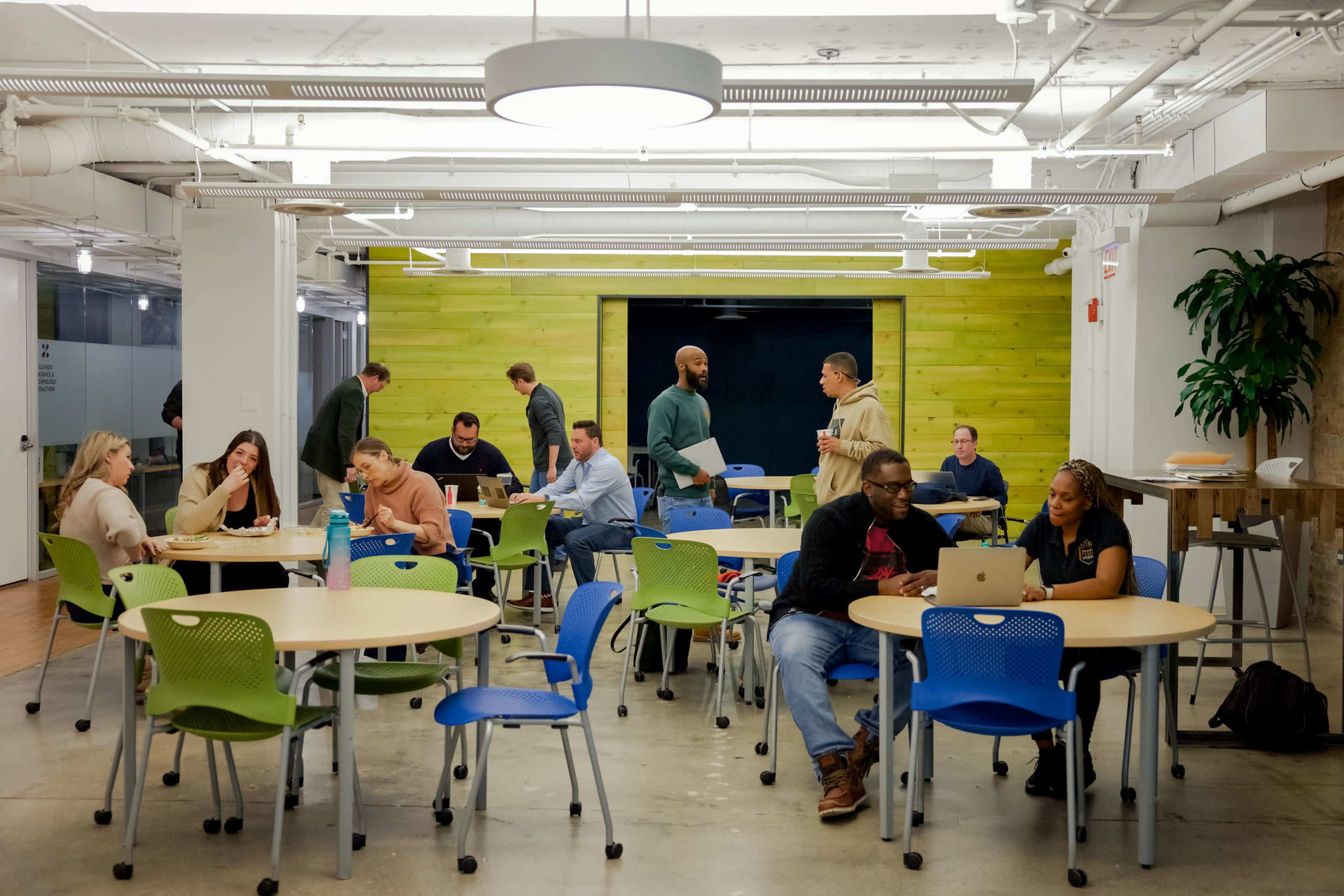 A diverse group of people work collaboratively at tables in a modern, well-lit workspace with green accents and an open design.