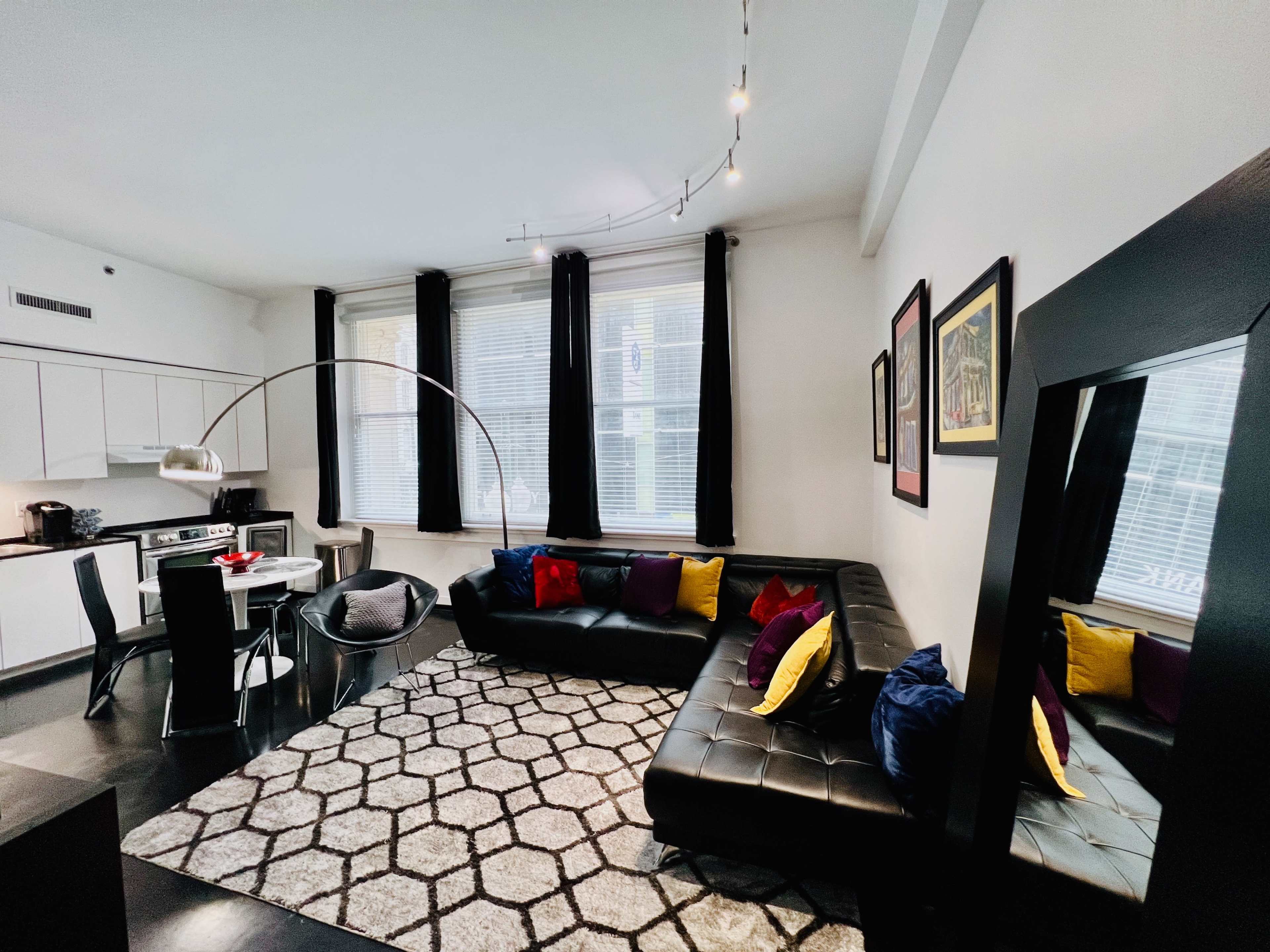 A modern living space features a black sectional sofa with colorful cushions, a circular dining table, and large windows adorned with black curtains.