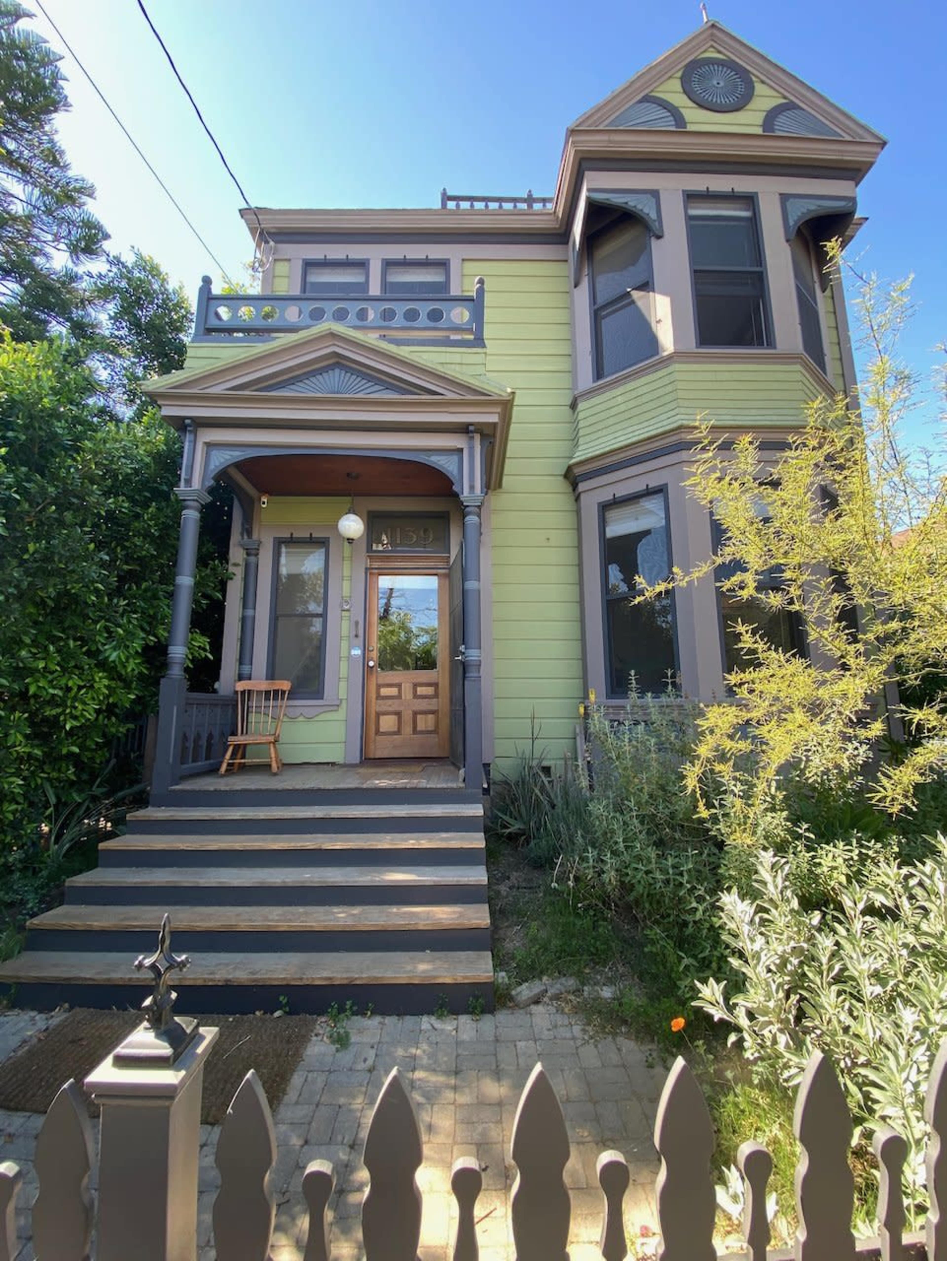 The image shows a two-story Victorian-style house with light green siding, a brown front door, and a porch supported by columns, surrounded by shrubs and a picket fence.
