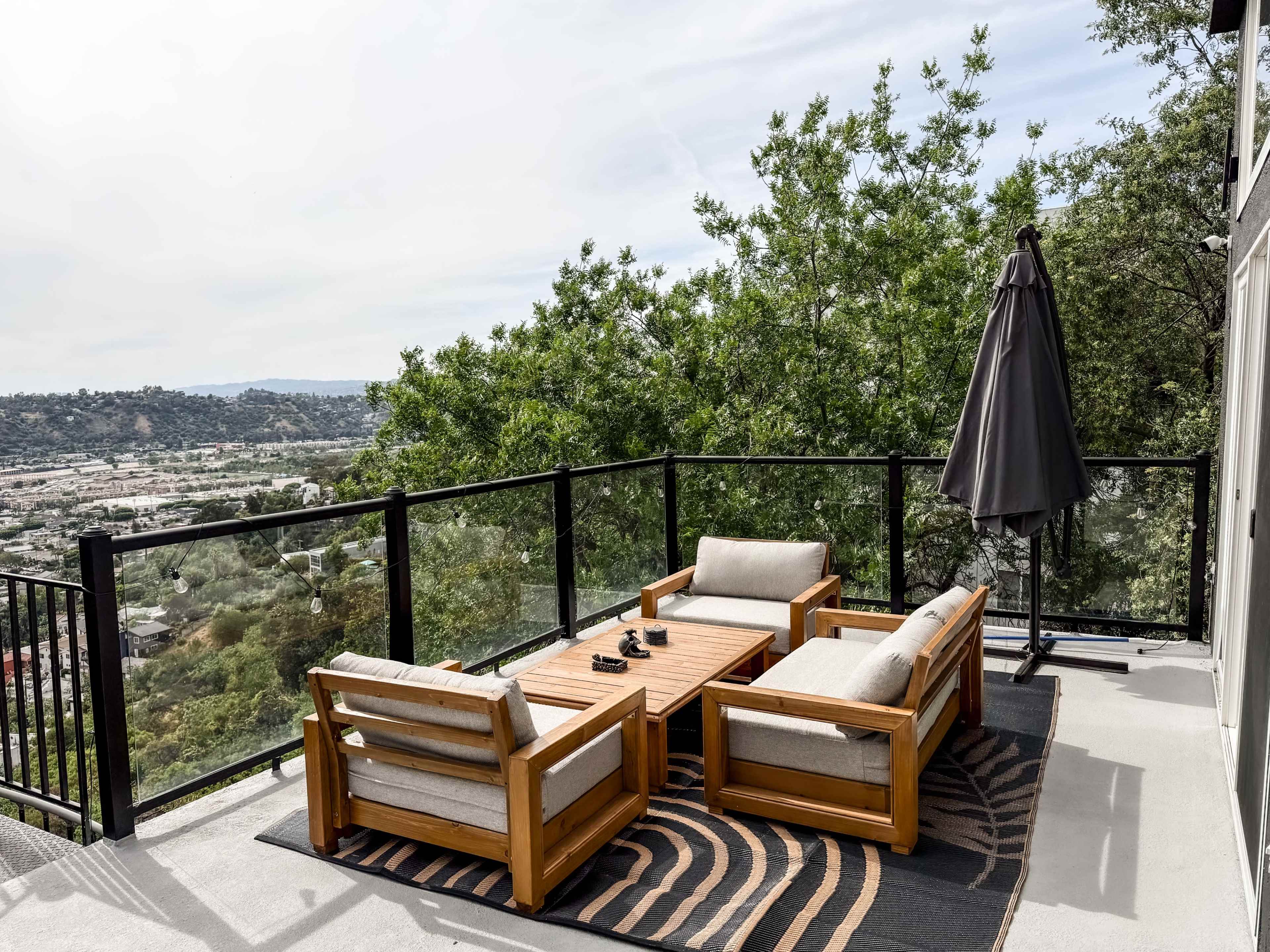 A wooden outdoor seating area with two chairs and a coffee table is set on a balcony overlooking a valley, surrounded by trees and featuring a patio umbrella.