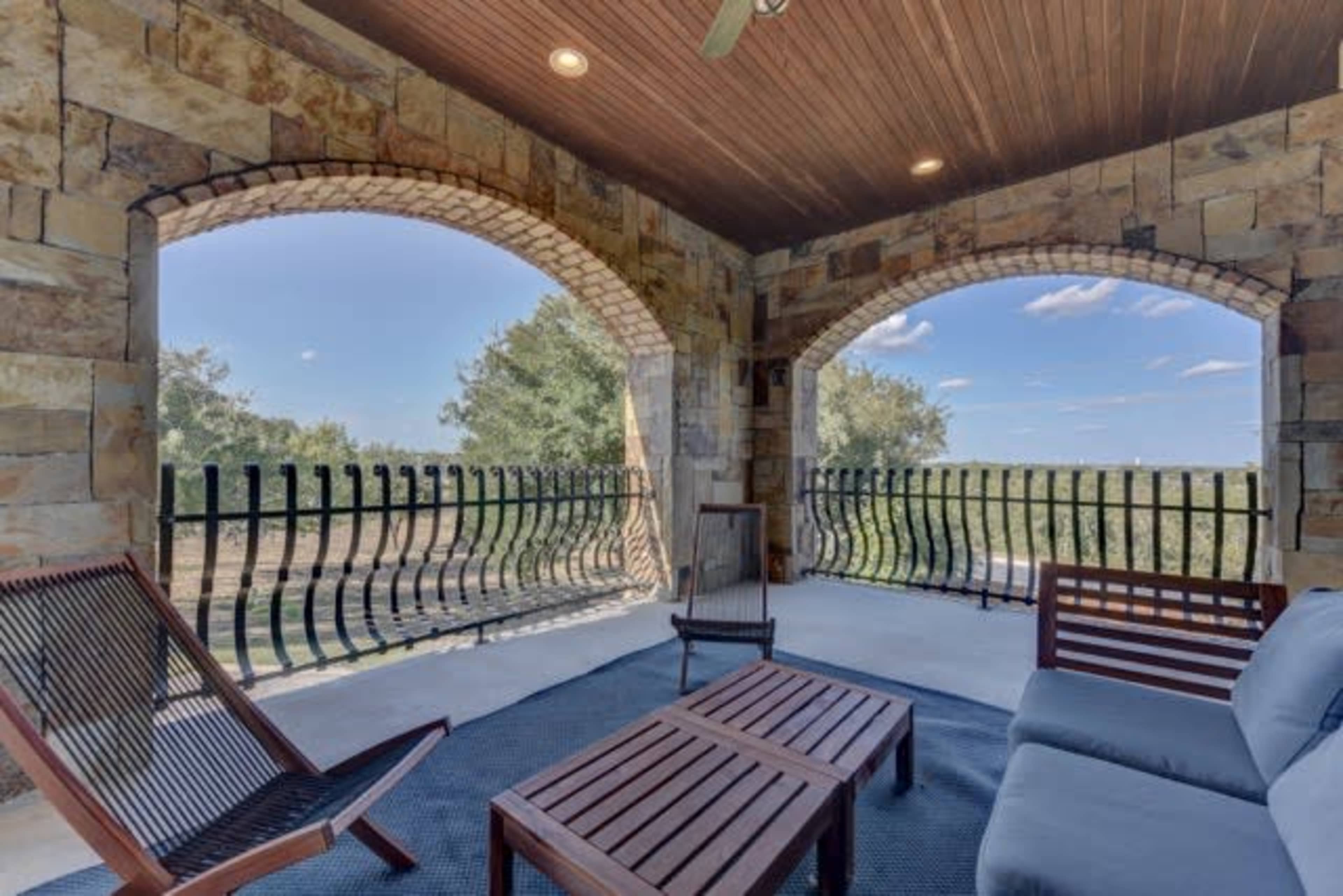 A covered patio with stone walls and arched openings overlooking a grassy landscape, furnished with wooden seating and a small table.