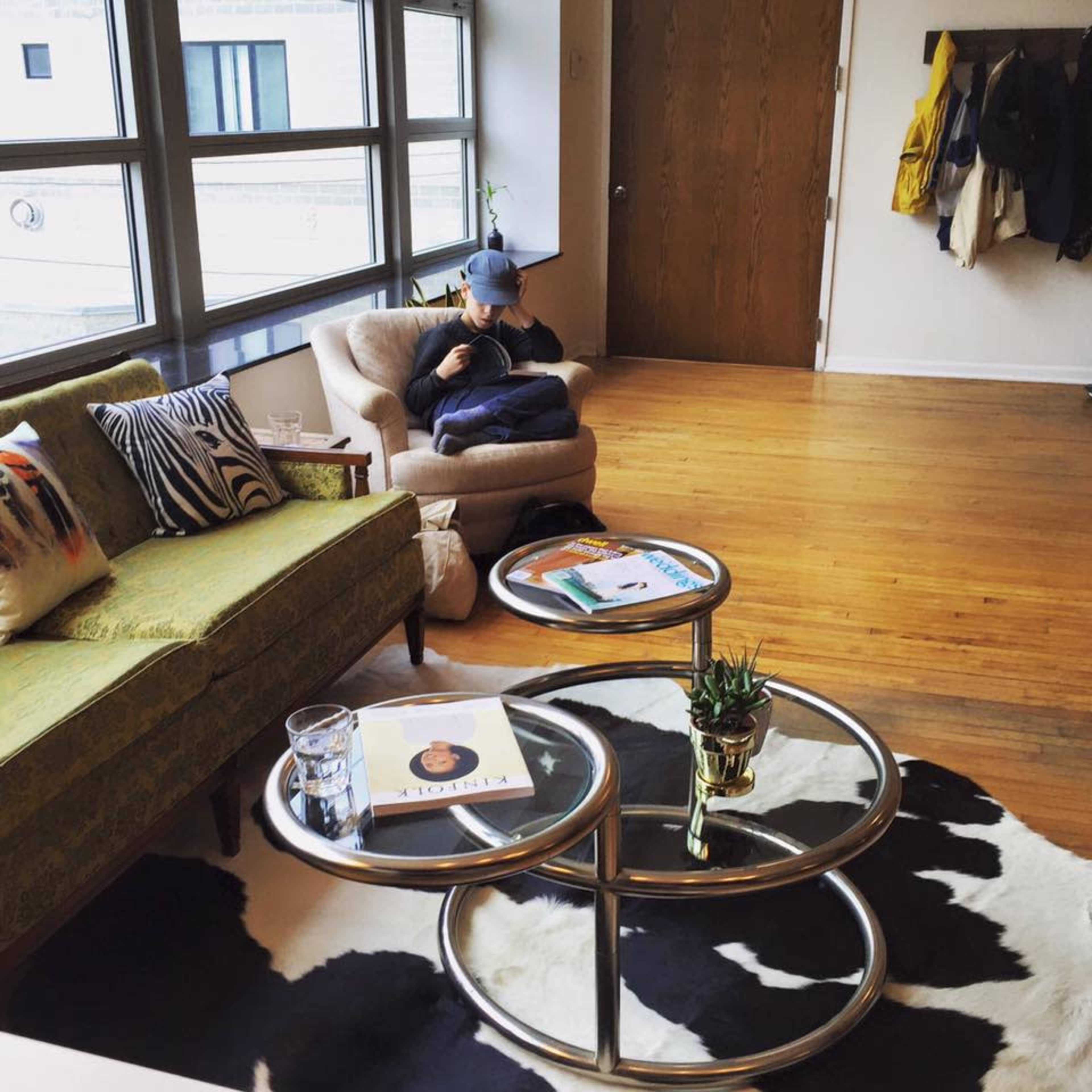 The image shows a cozy living room with a green sofa, a person seated on an armchair, and a circular coffee table on a cowhide rug.