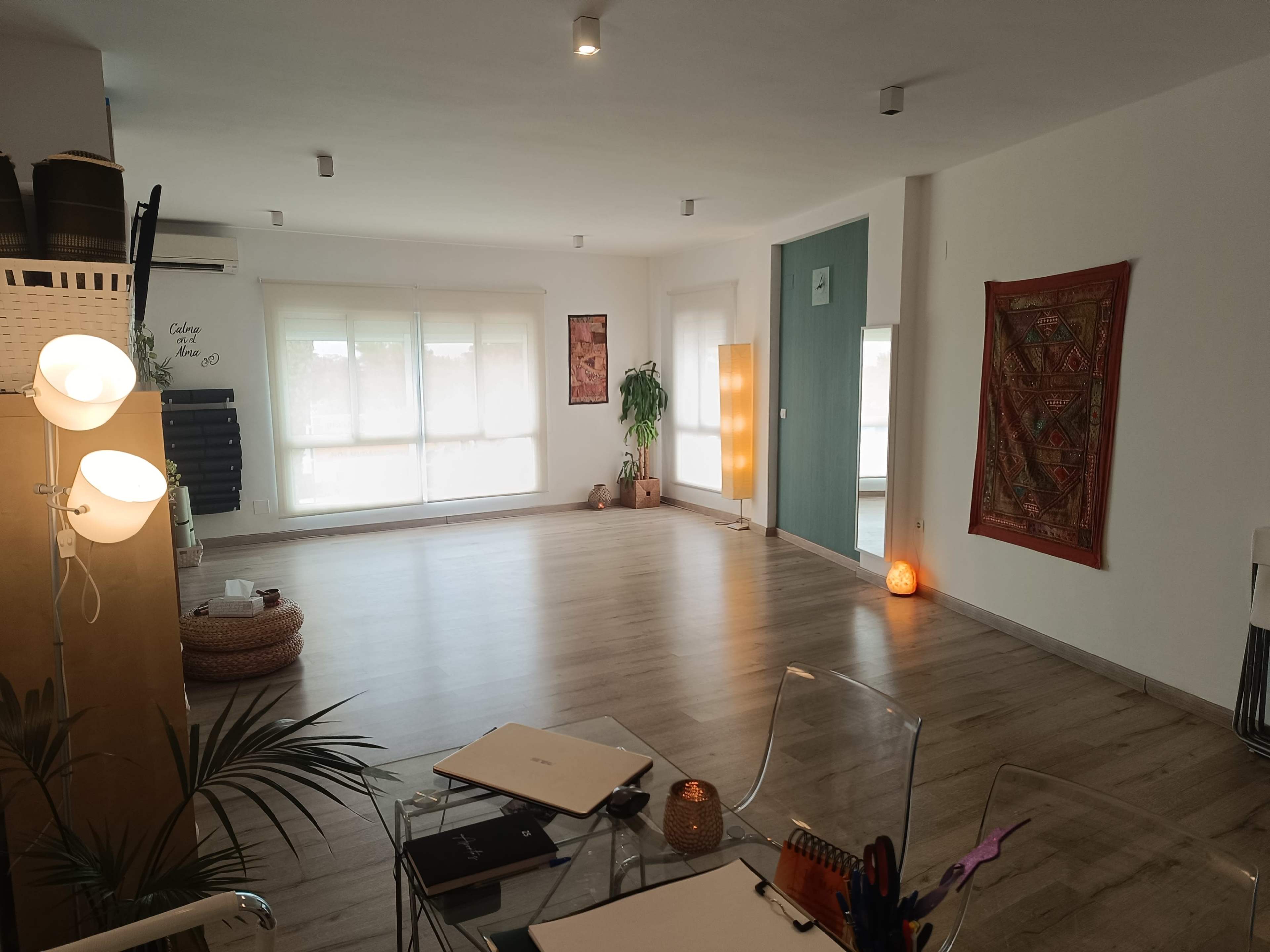 The image shows a spacious, well-lit room with wooden floors, large windows, a potted plant, and various decor items on the walls.
