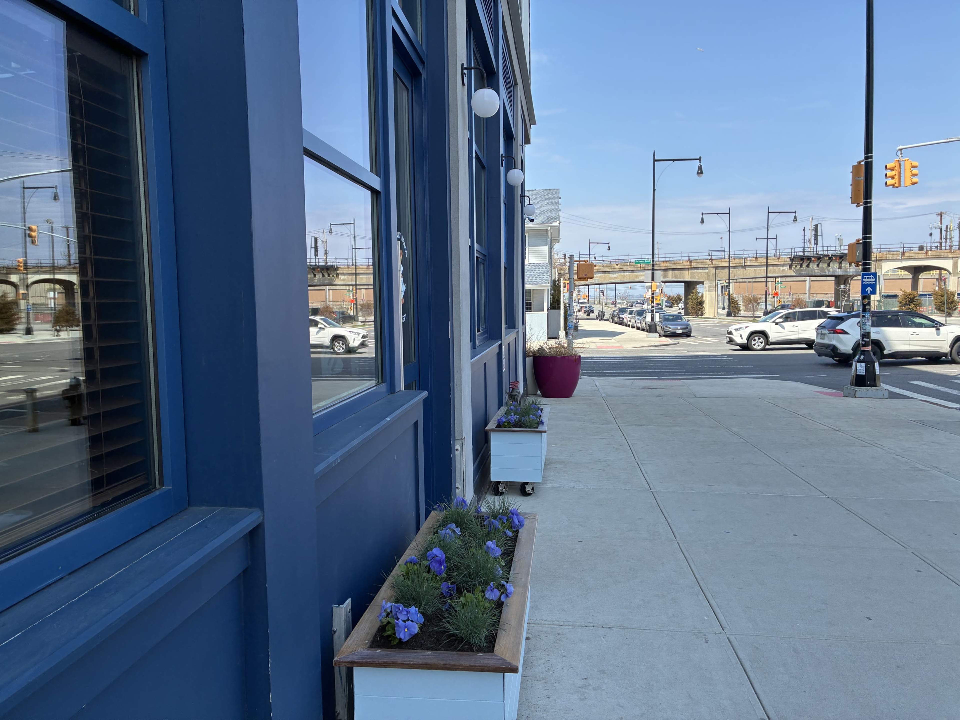 The image shows a sidewalk alongside a building with blue walls and planters containing flowers, adjacent to a street with passing cars and traffic signs.