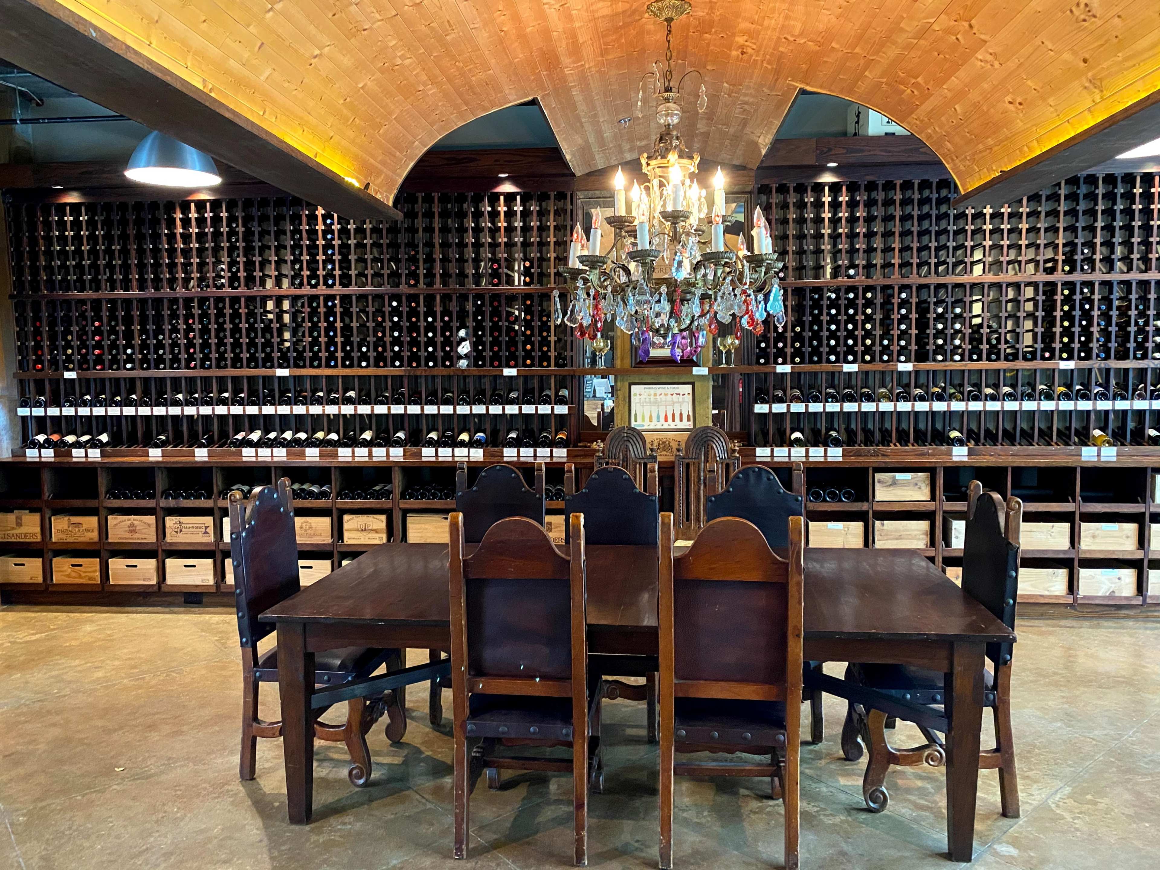 A large wooden dining table with sturdy chairs is set in front of a wall filled with neatly arranged wine bottles and a chandelier overhead.