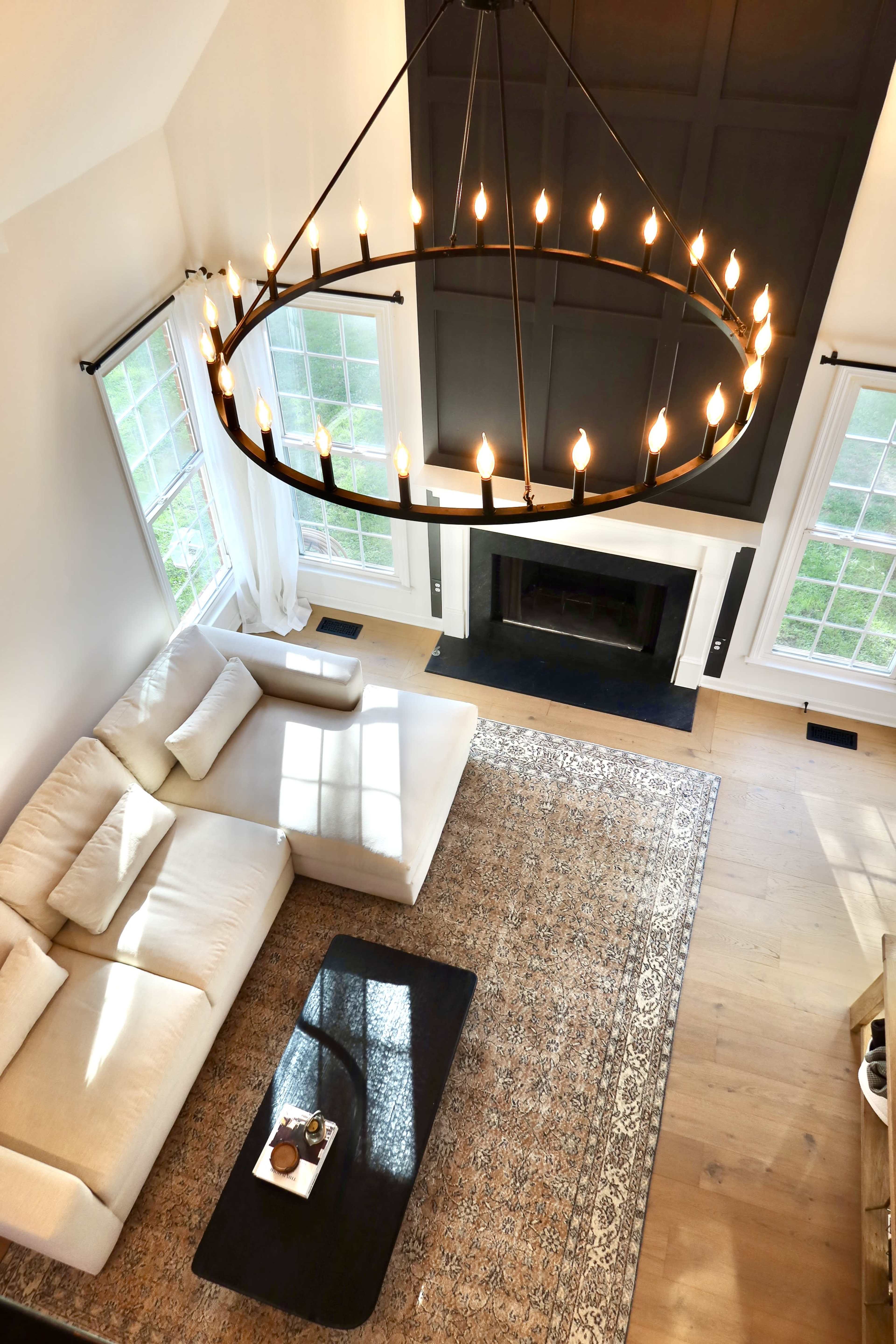 The image shows a spacious living room with a large sectional sofa, a circular chandelier, and a coffee table on a patterned rug, all beneath a high ceiling with large windows.