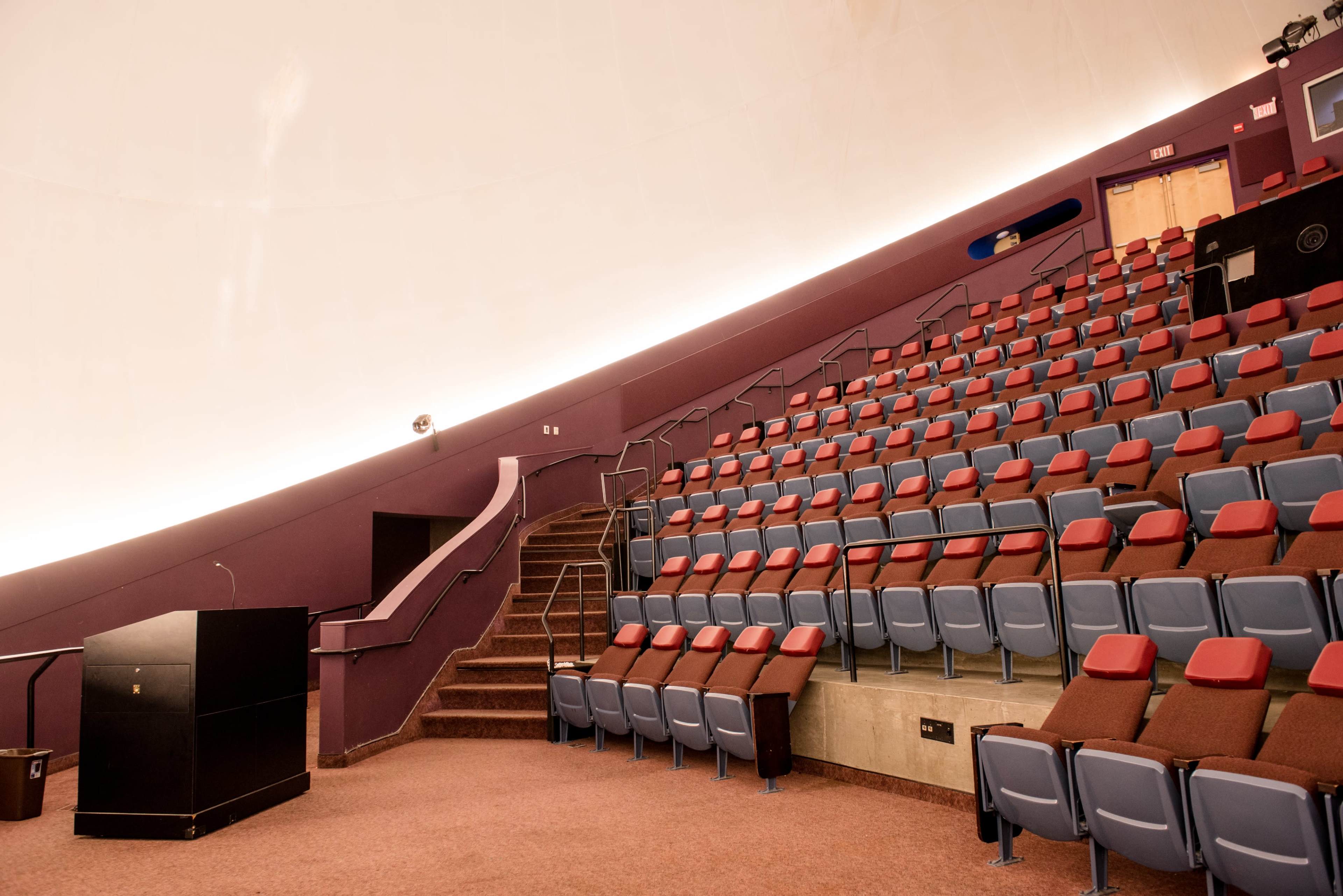 The image shows an empty auditorium with tiered seating, a podium on the stage, and a domed ceiling.