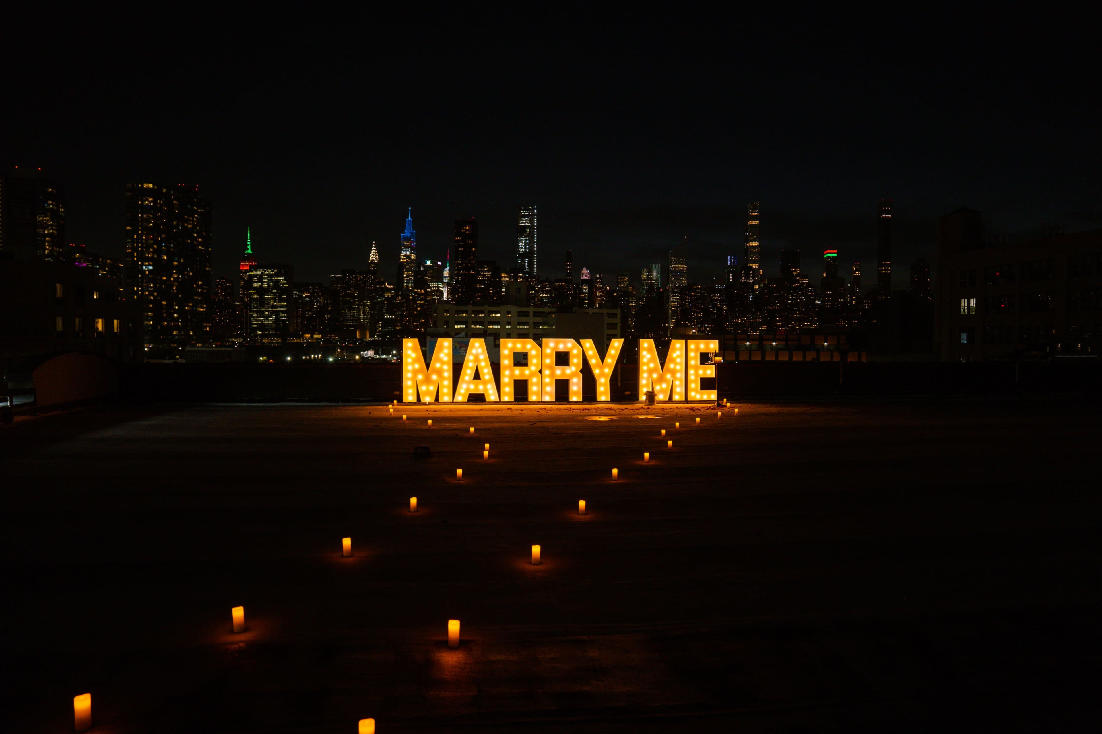 The scene features large illuminated letters spelling "MARRY ME" on a rooftop, surrounded by candles, set against a nighttime skyline of a city.