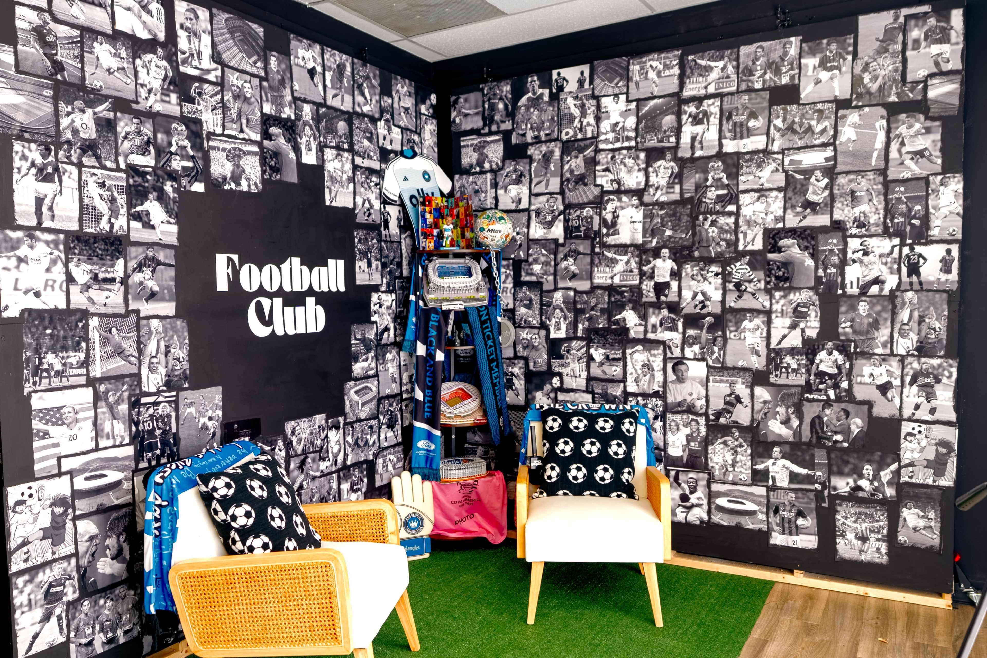 The image features a room decorated with a wall covered in black and white photographs related to football, alongside two white chairs and artificial grass on the floor.