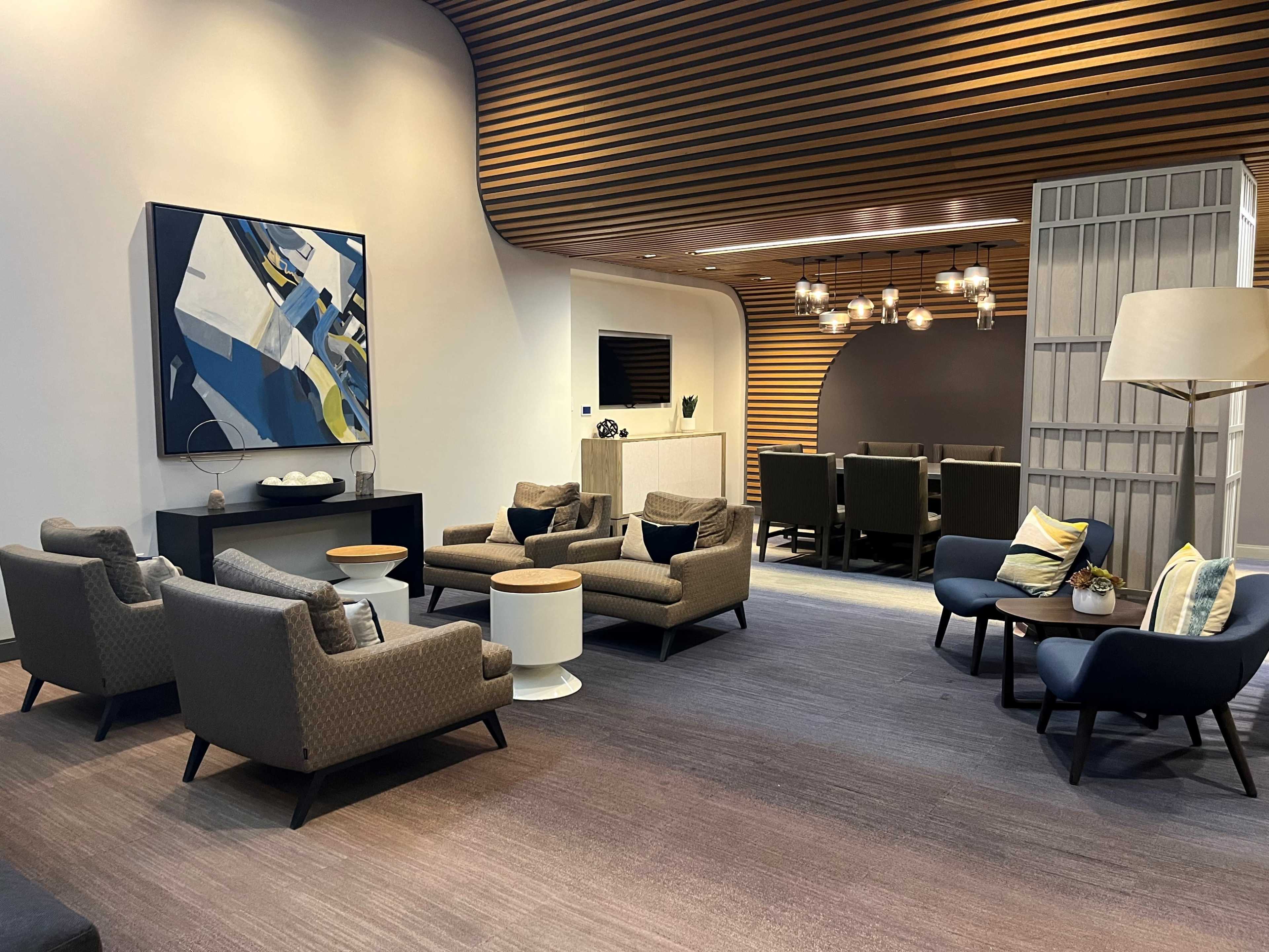 The image shows a modern lounge area featuring several armchairs, small tables, and a large abstract painting on the wall, with a dining area visible in the background.