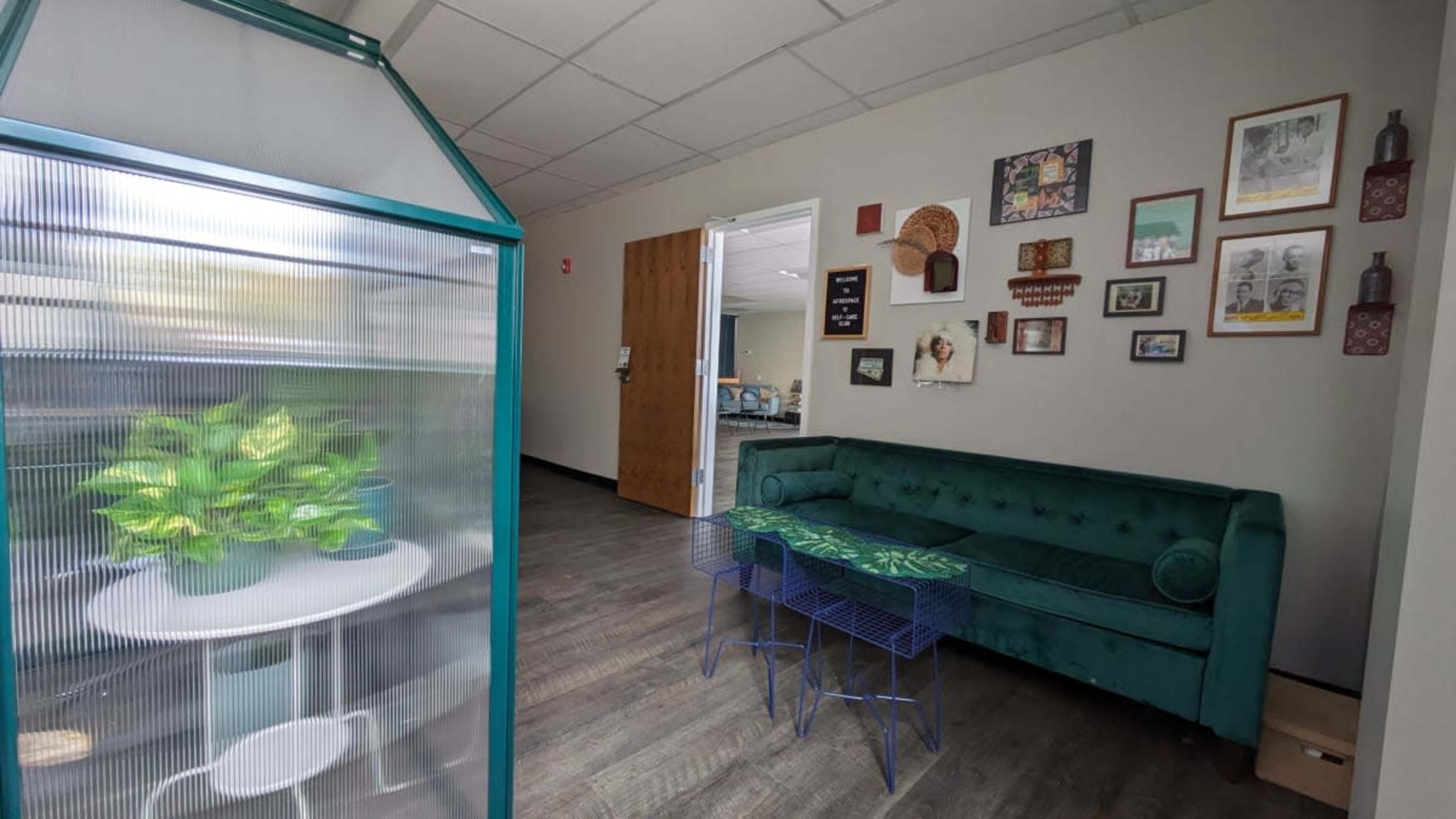A well-lit room featuring a green couch, a small round table, blue metal side tables, and a greenhouse with plants.