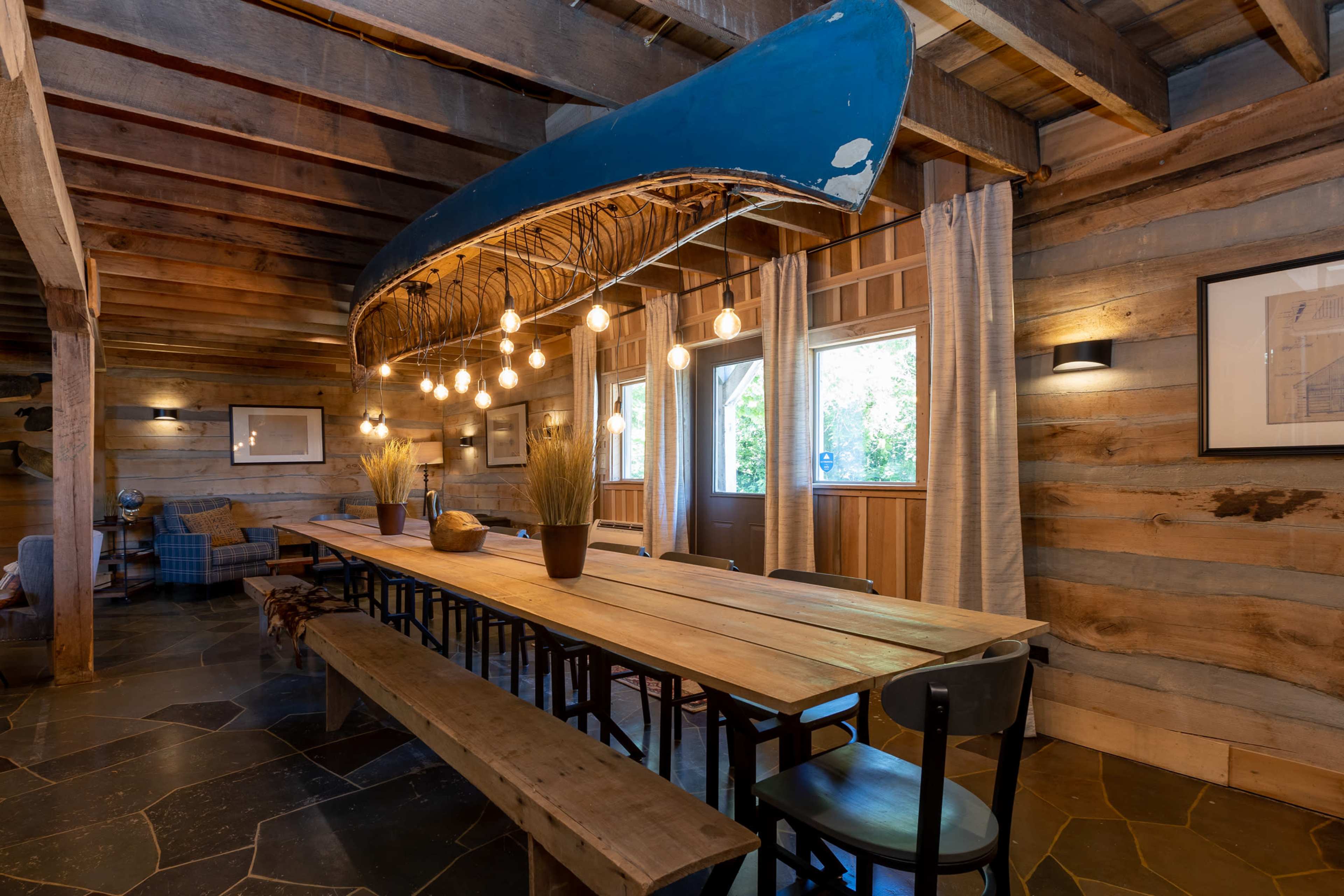 A rustic dining area features a long wooden table with benches, illuminated by pendant lights suspended from a canoe above.