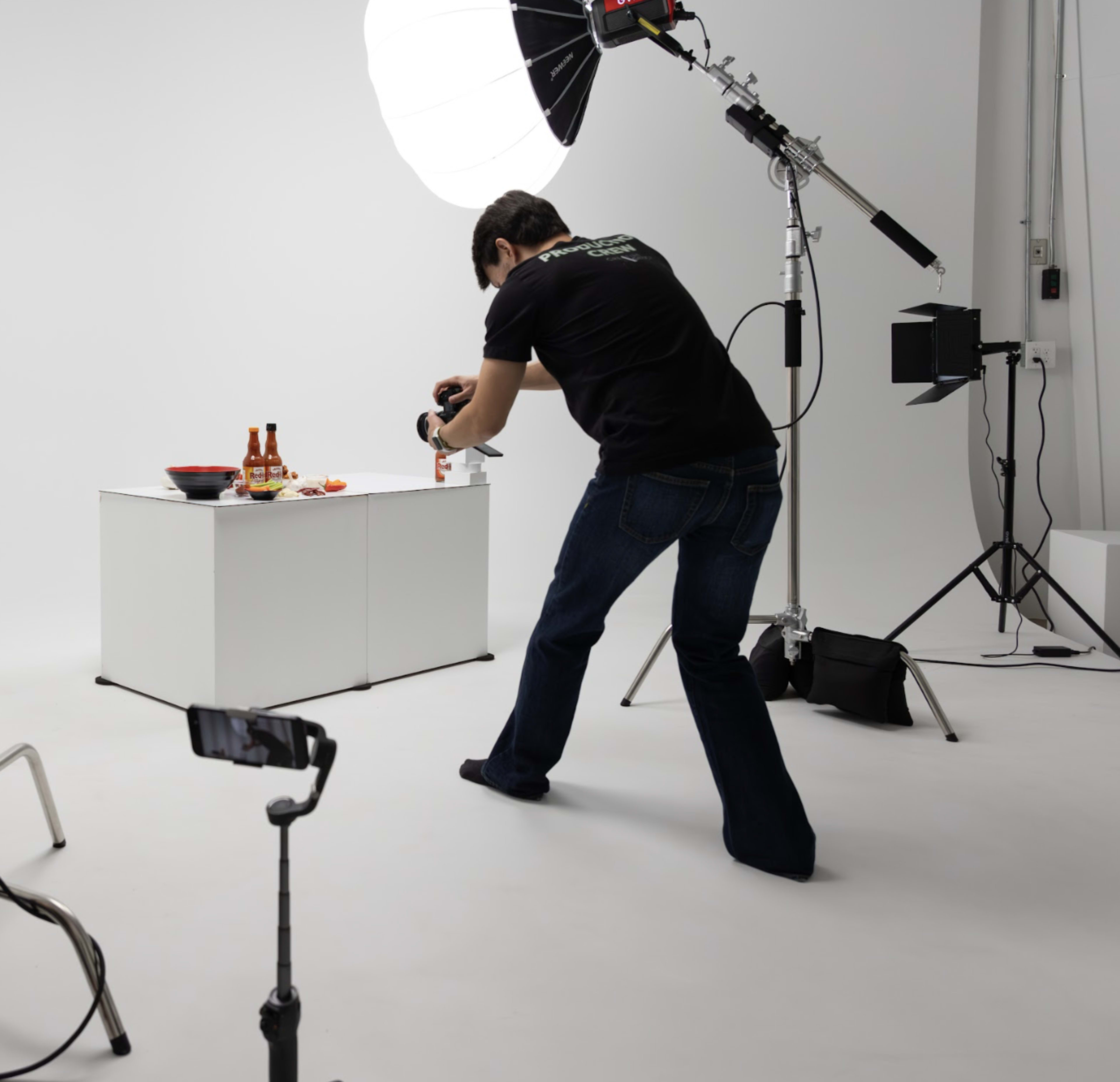 A photographer wearing a black shirt captures an image of food on a table in a studio setting with multiple lighting sources.