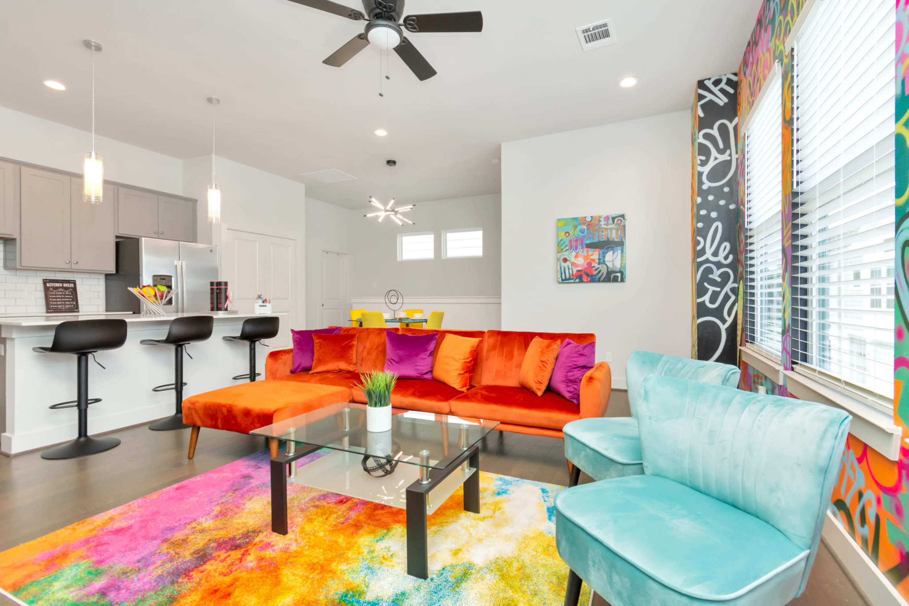 The image shows a brightly decorated living area with a colorful sofa, glass coffee table, and stylish chairs, adjacent to a modern kitchen.