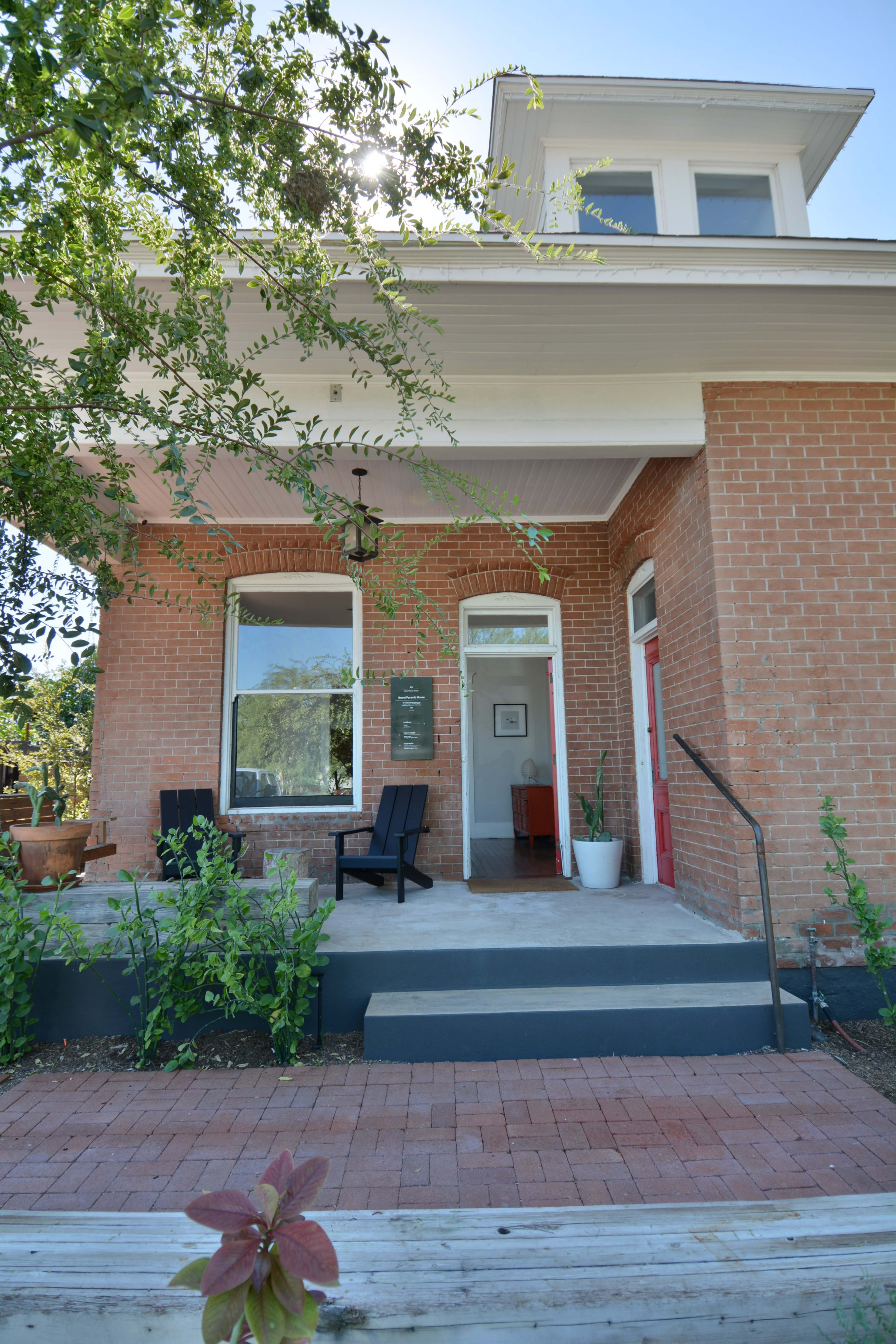 A brick house with a porch, featuring two front doors, a potted plant, and chairs on the patio.