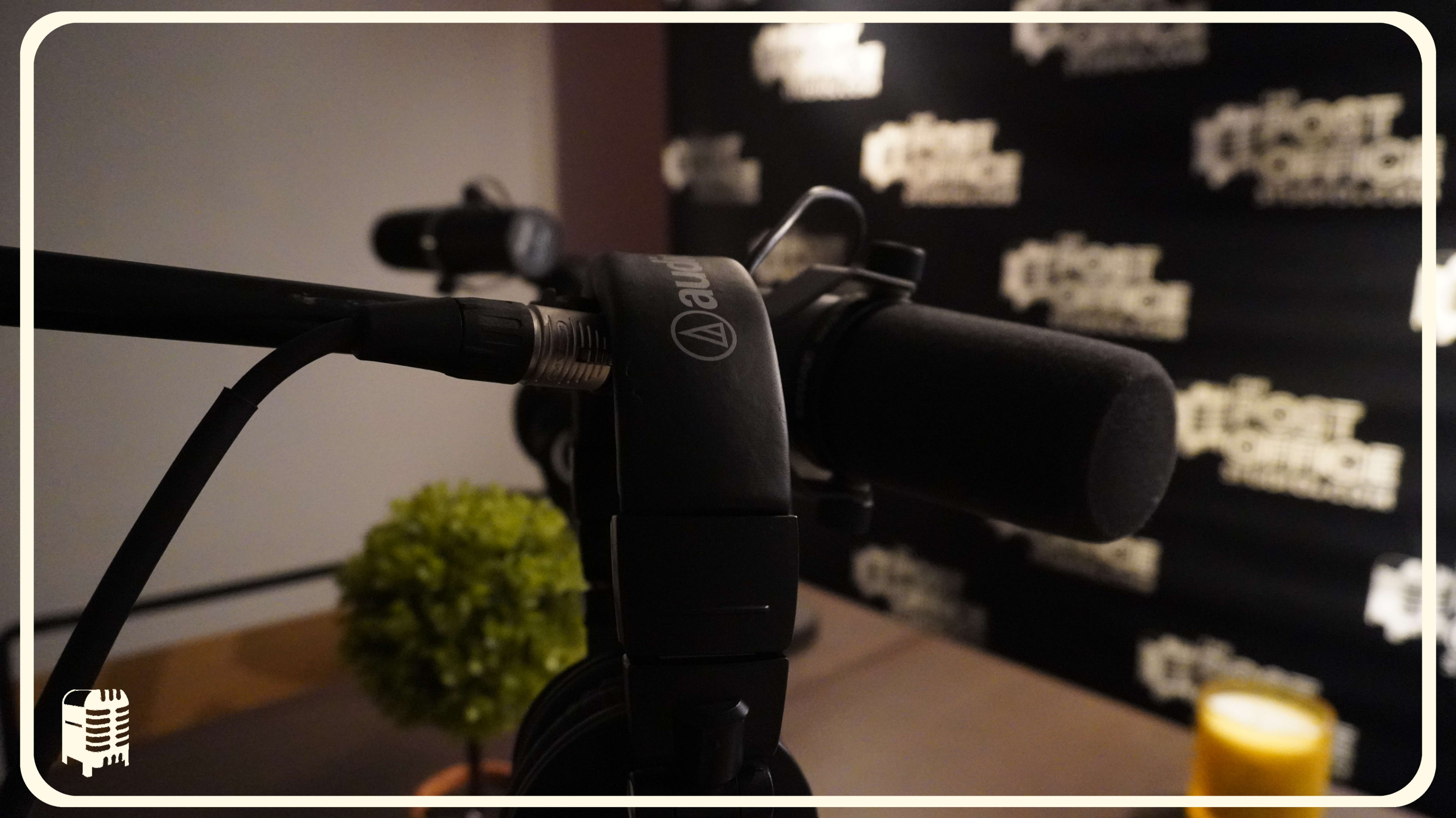 A close-up of a microphone with a blurred background featuring a logoed wall and a small potted plant on a table.