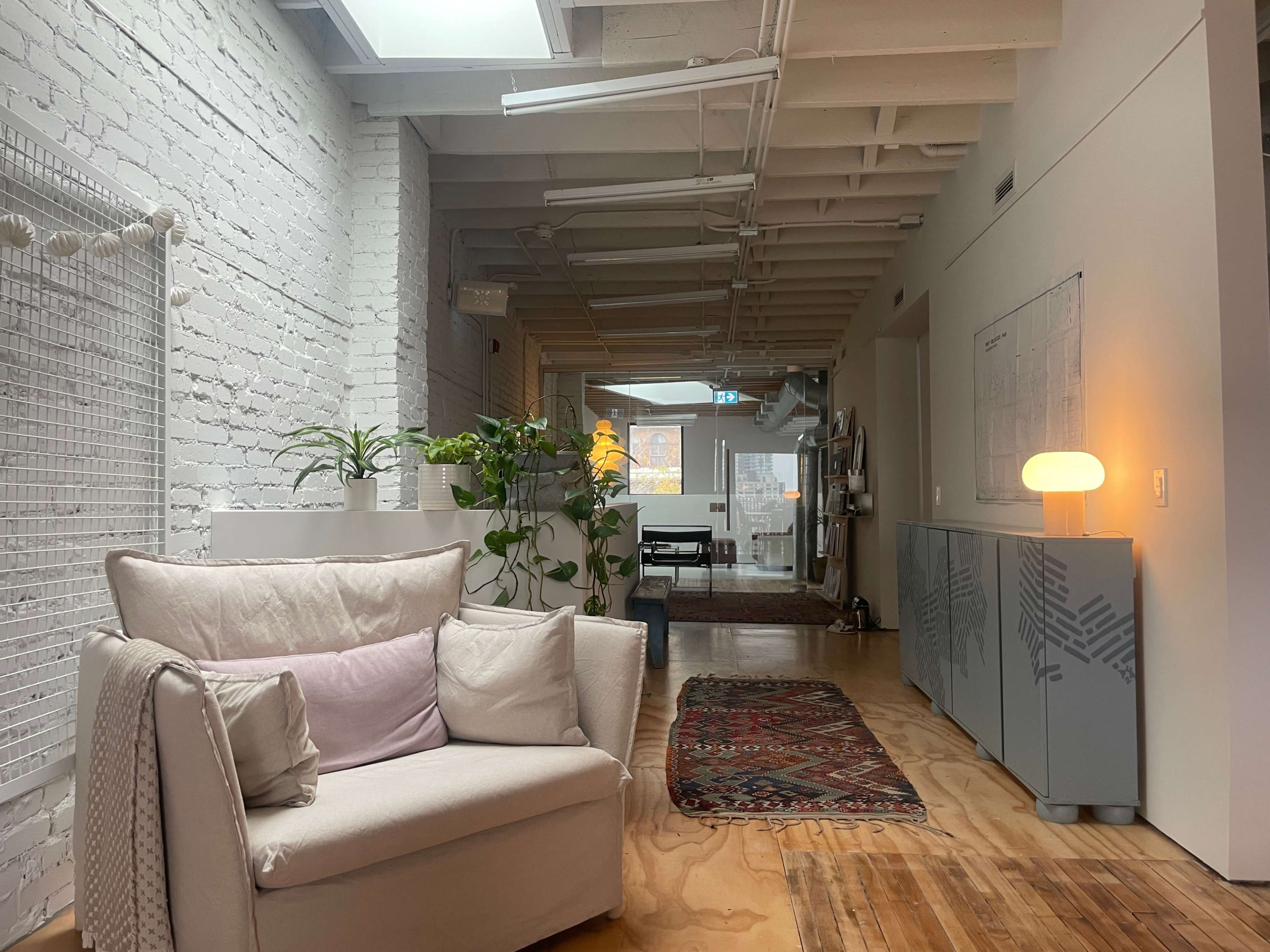 The image shows a spacious, modern living area with a white brick wall, wooden flooring, a light-colored sofa, and decorative plants, leading to a well-lit space with a rug and furniture in the background.