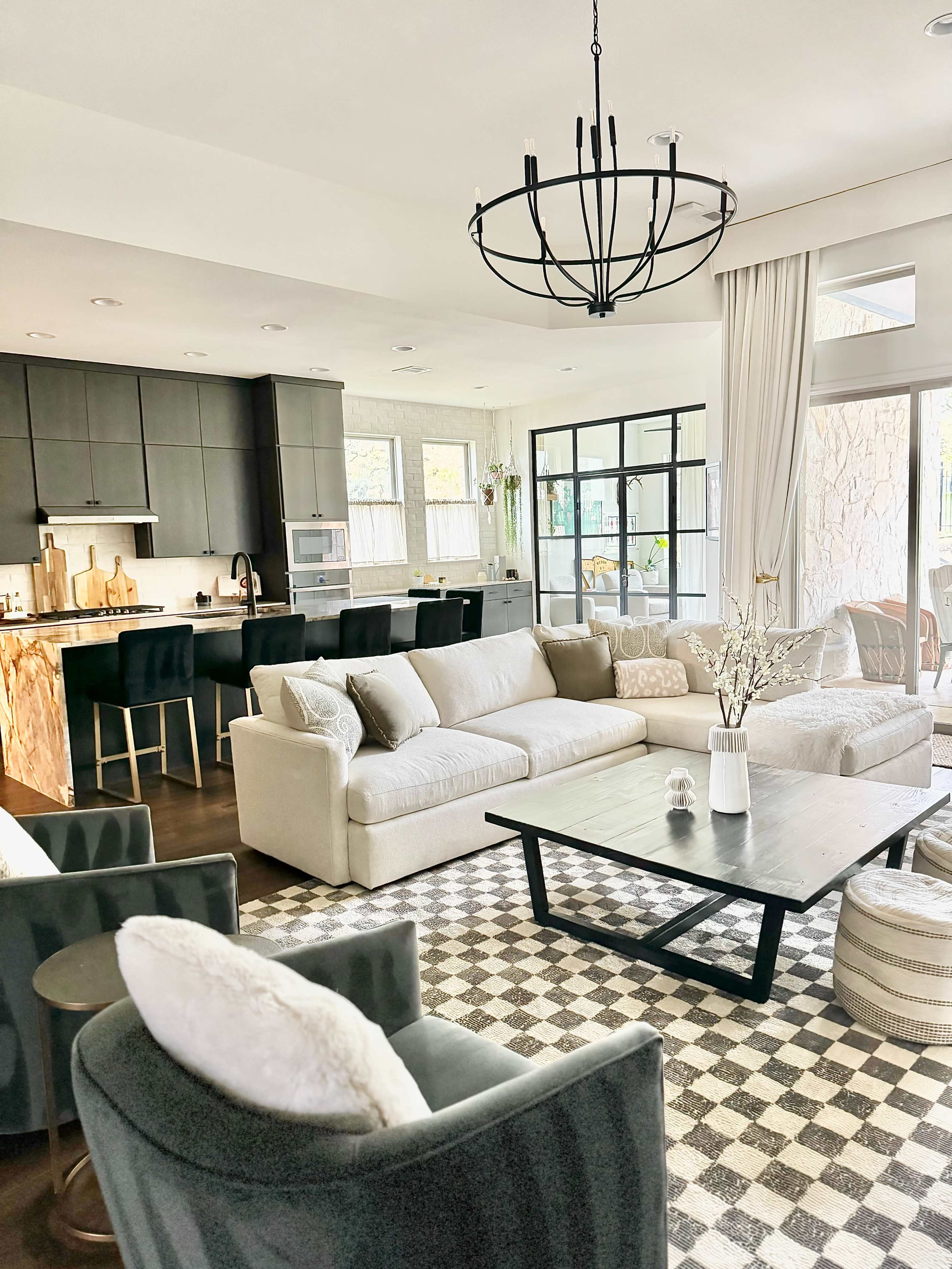 The image shows a modern living room with a large sectional sofa, a coffee table, and a kitchen area in the background, featuring a chandelier and a checkered floor pattern.