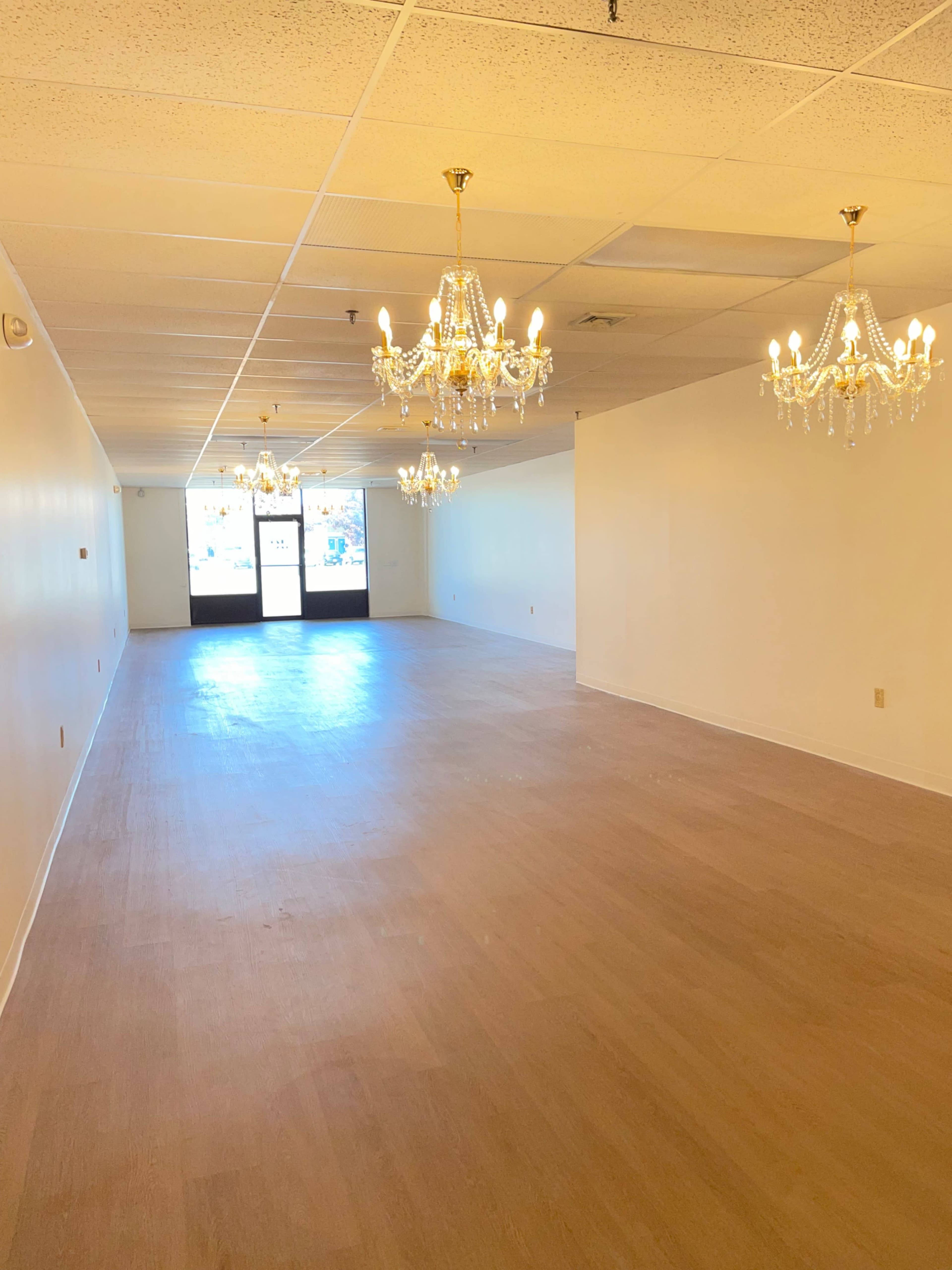 The image shows a spacious, empty interior room with wooden flooring and several chandeliers hanging from the ceiling.