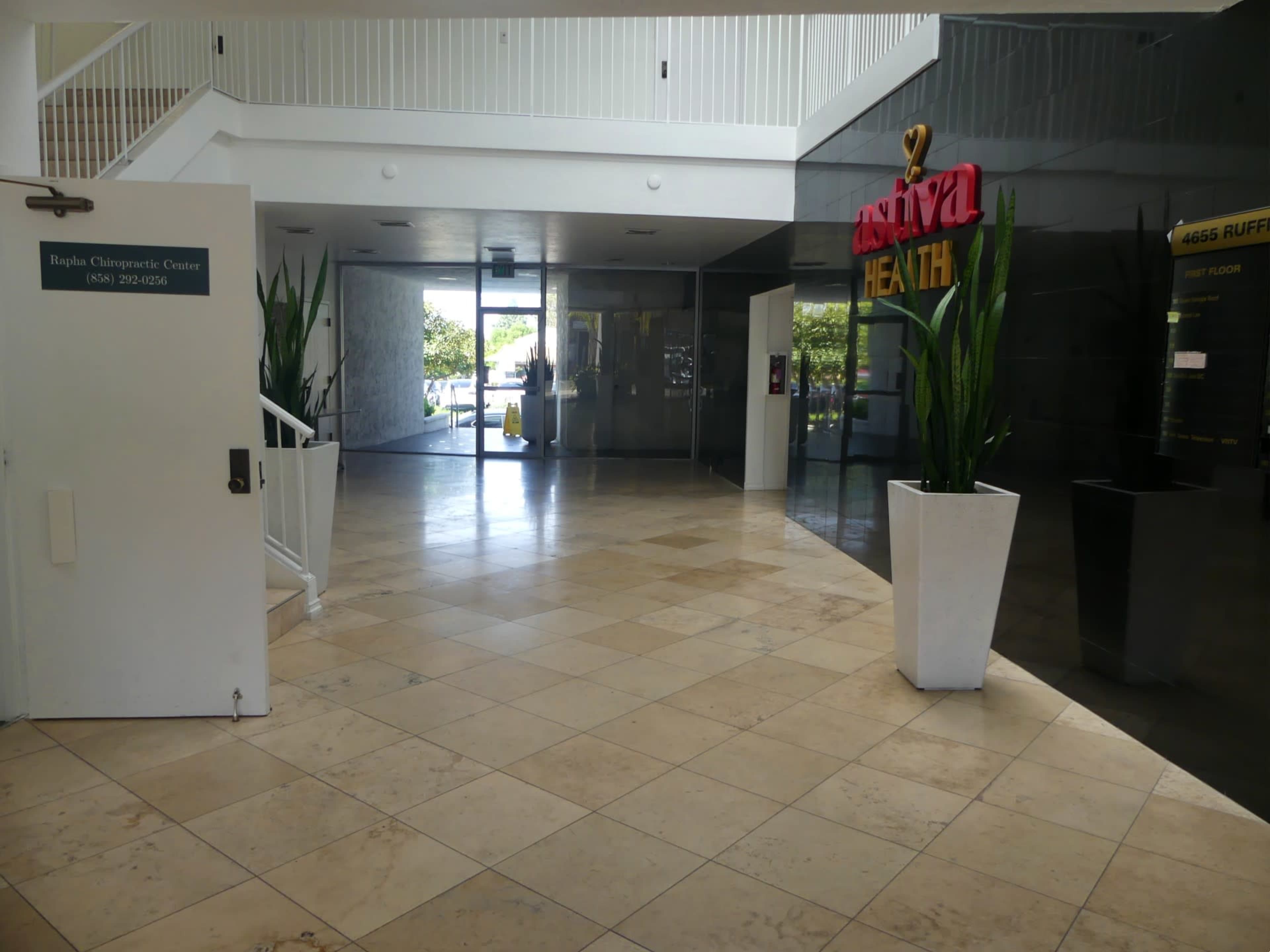 A spacious lobby area with tiled floors, featuring a reception desk and large potted plants, leading to a glass-walled entrance labeled "Riviva Health."