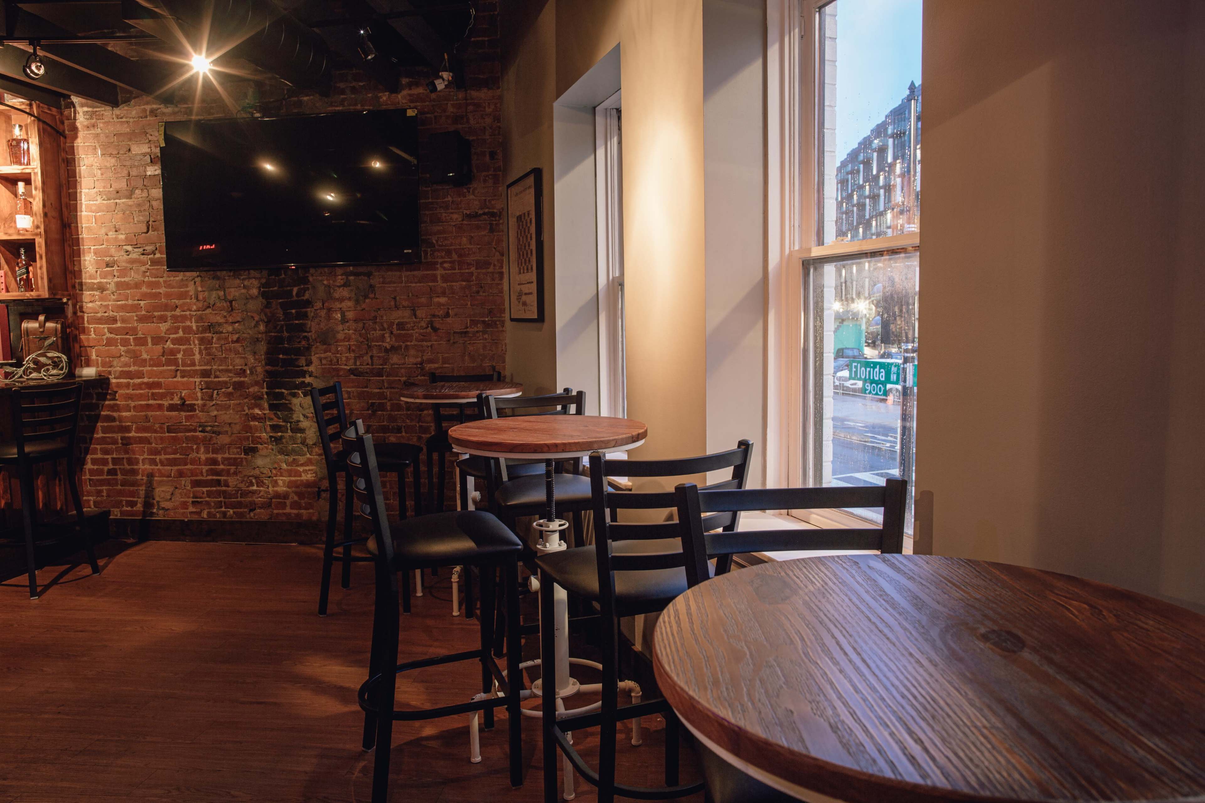 A cozy bar setting with wooden tables and chairs, a brick wall, and a large window facing a street.