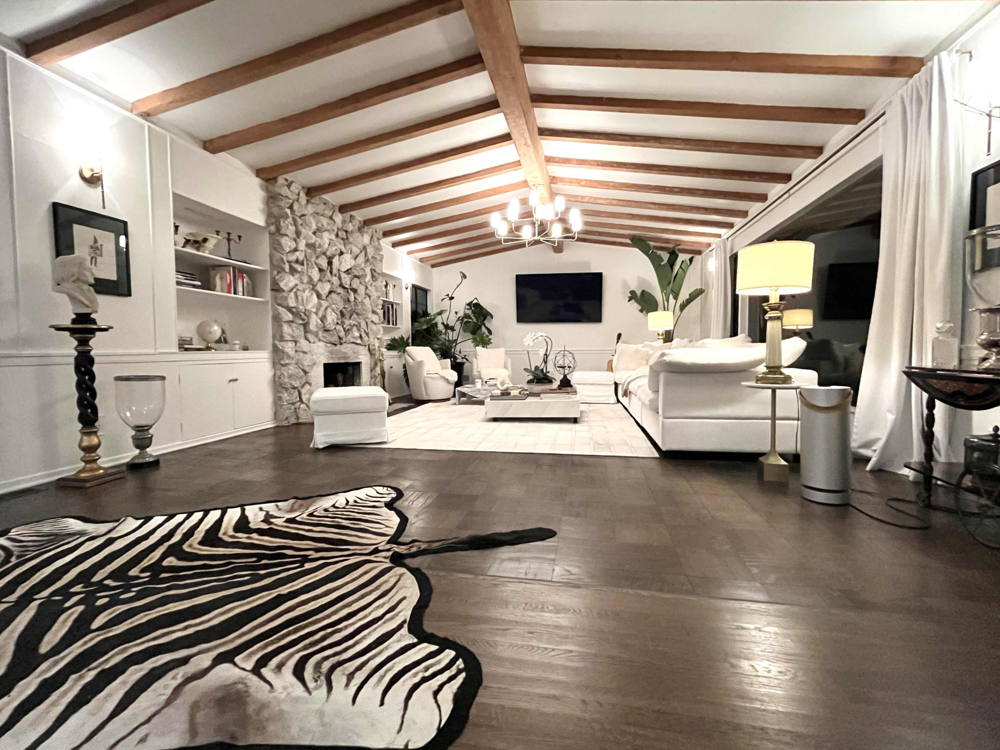 The image features a spacious living room with exposed wooden beams, a stone accent wall, and modern furnishings surrounded by indoor plants.