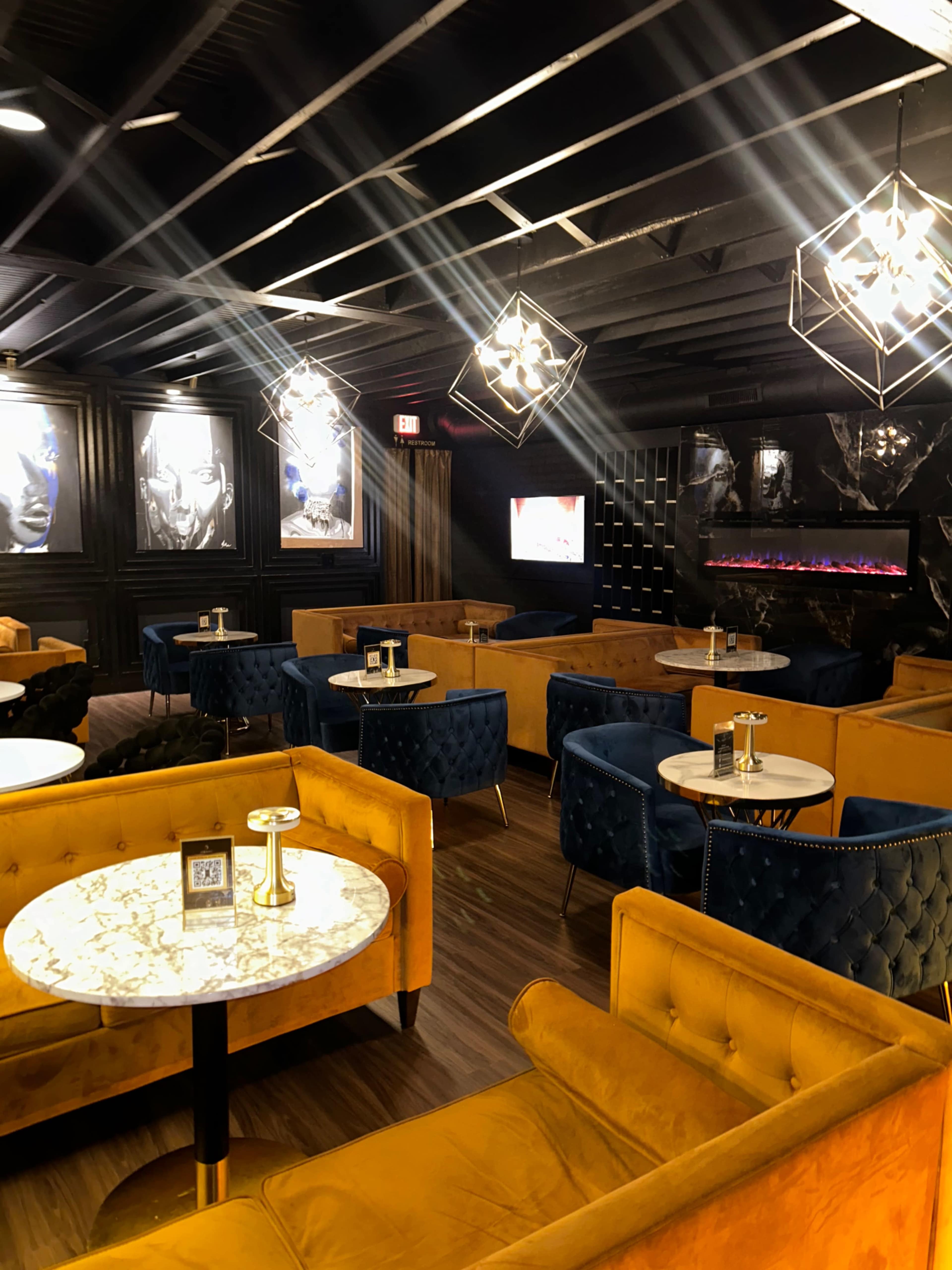 The image shows a stylish lounge area with orange and blue upholstered seating, round marble-topped tables, and modern light fixtures, set against a dark, textured wall.